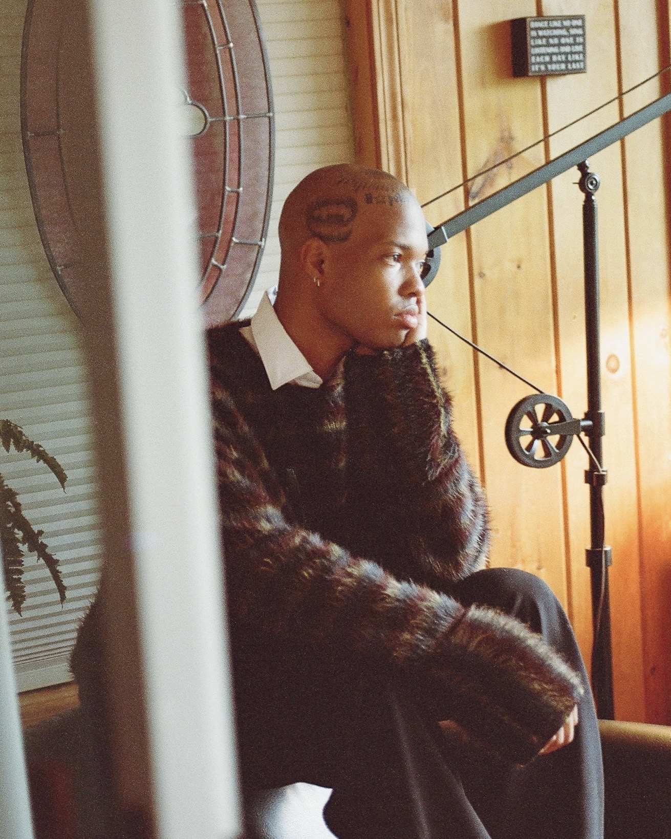 Person with braided hair sitting indoors, looking to the side, with wooden walls and a microphone stand nearby.