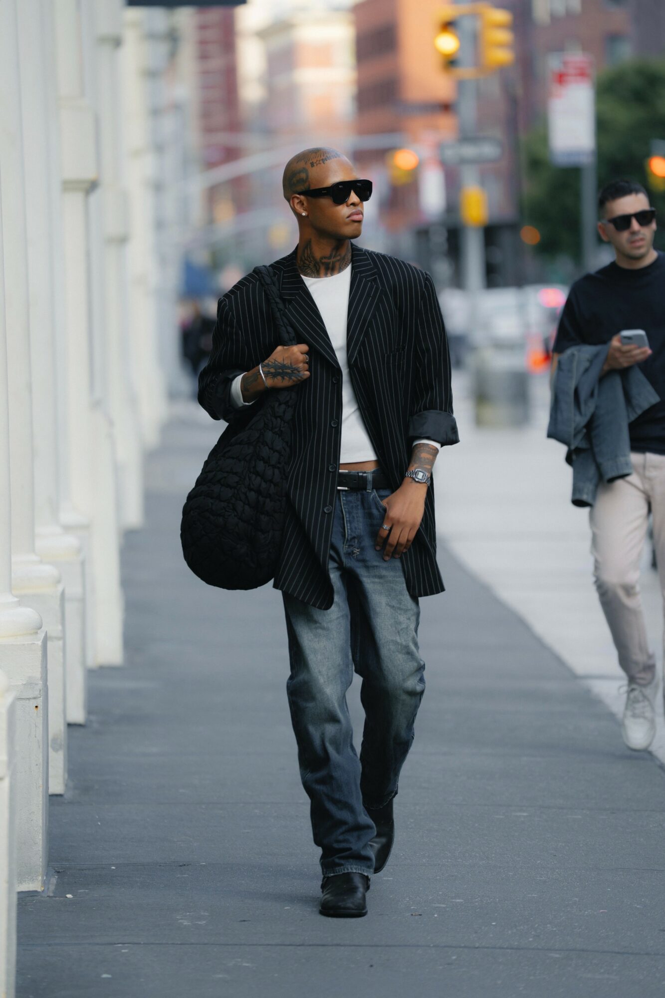Man walking on city sidewalk wearing sunglasses, black coat, white shirt, and jeans, carrying a large bag, with another person in background.