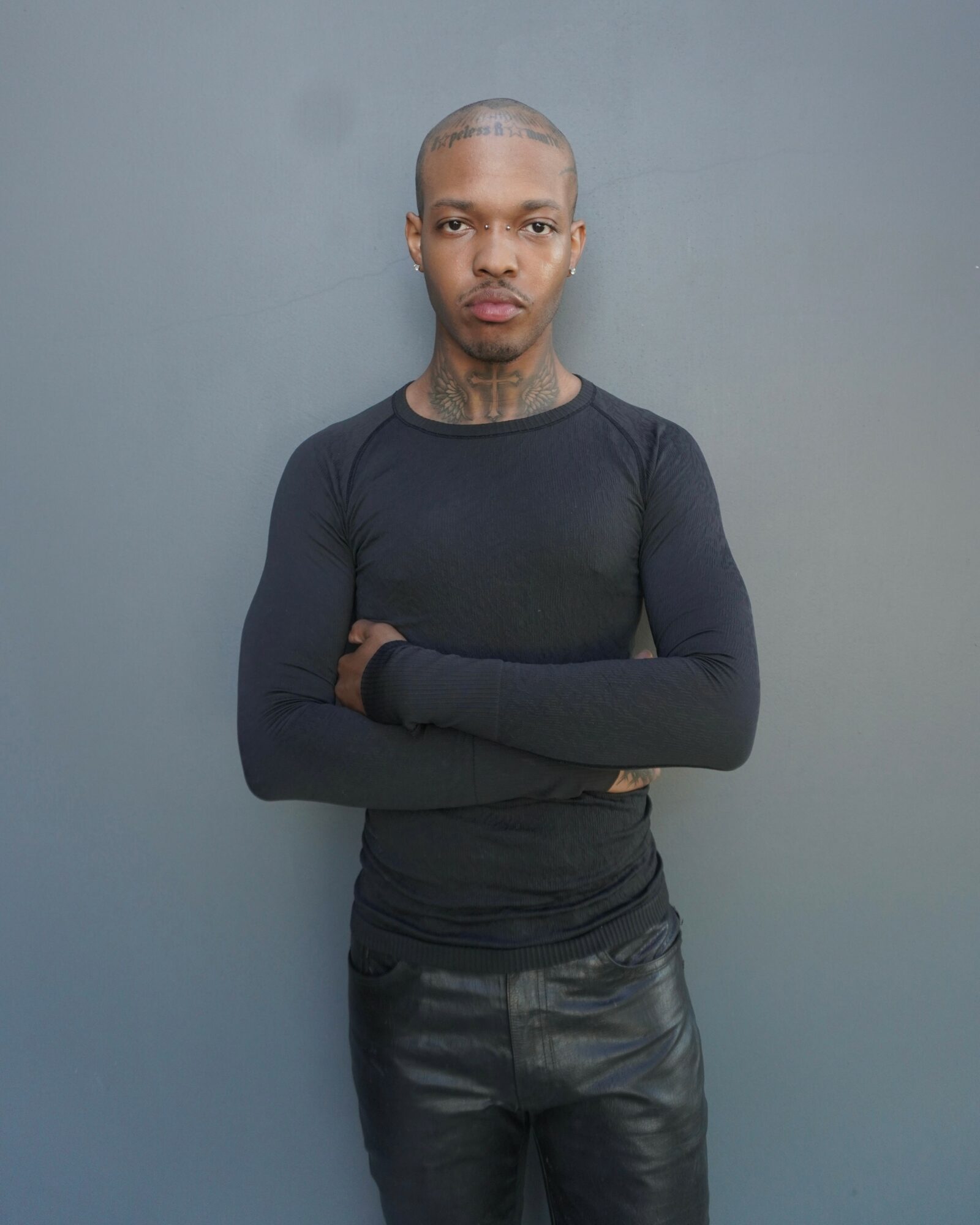 Person with short hair and tattoos, wearing a black long-sleeve shirt and leather pants, standing against a gray wall.