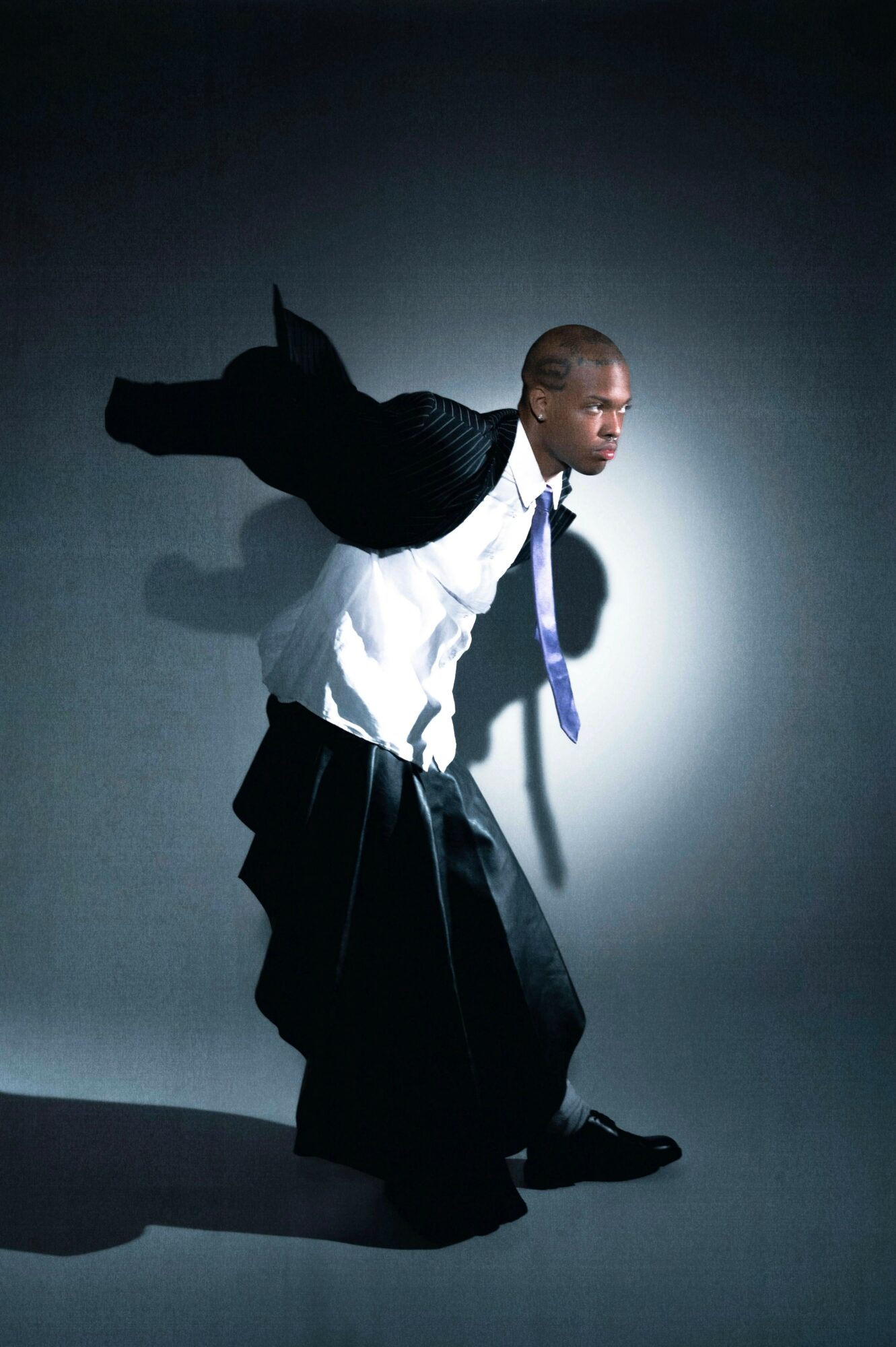 Person in traditional clothing with a jacket draped over shoulders, standing against a dark background, looking to the side.