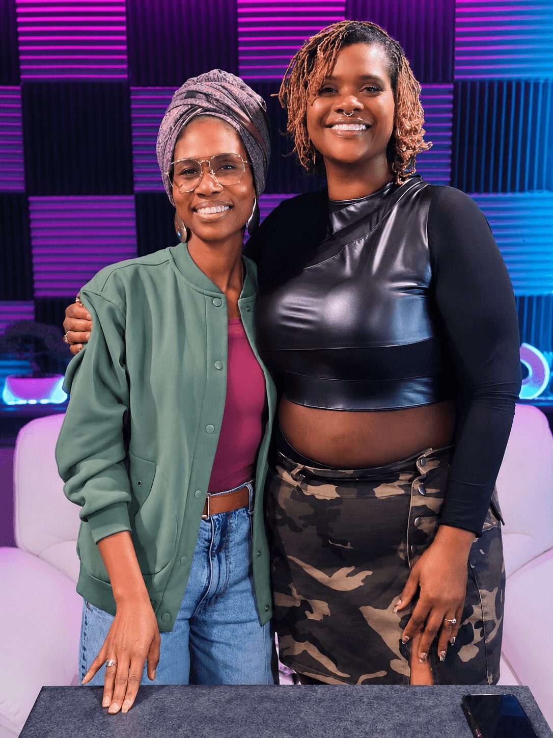 Two women smiling, one with glasses and a headscarf, the other with curly hair, standing in a colorful studio.