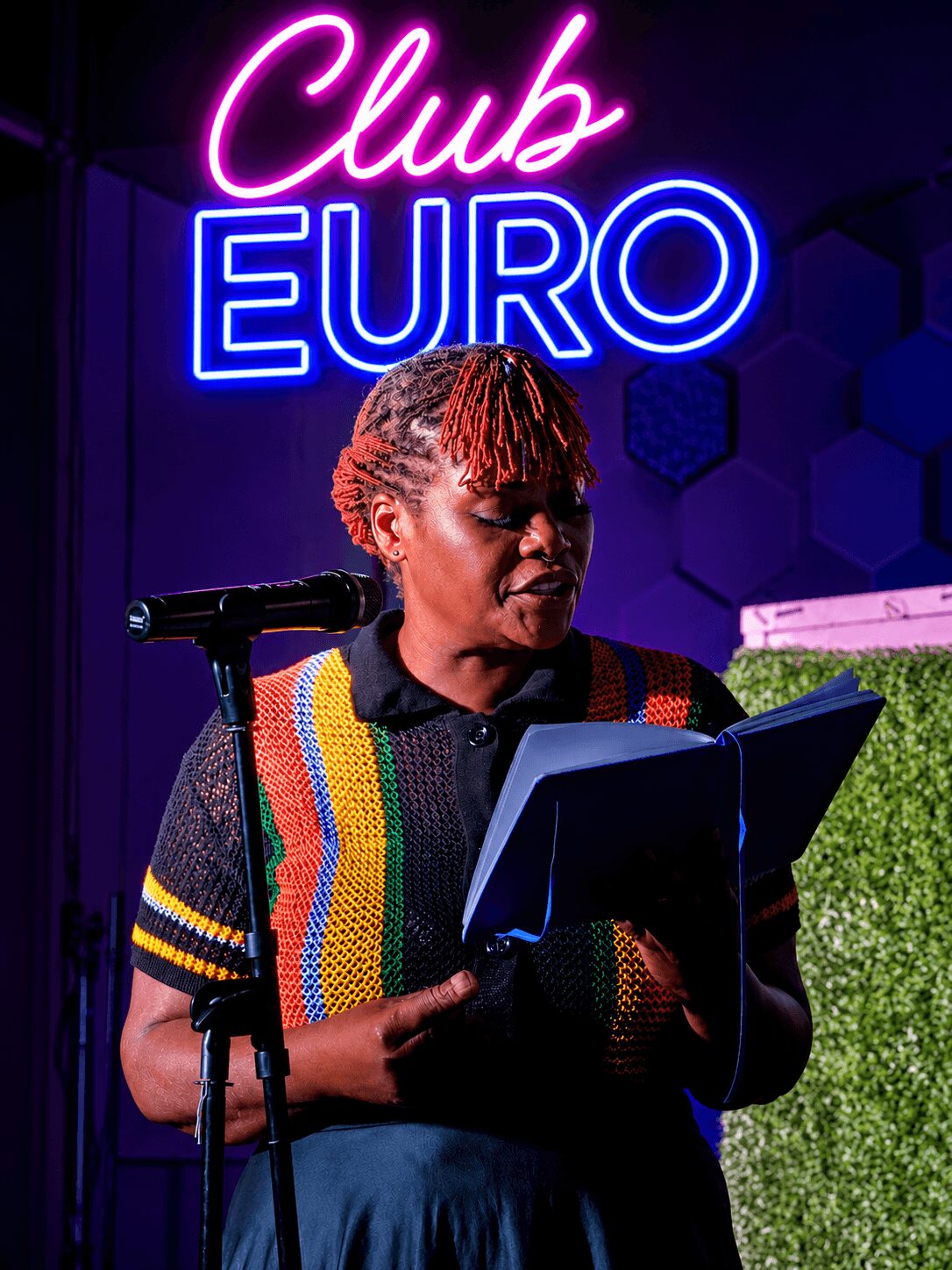 Woman reading from a tablet on stage with a microphone, neon sign reads 'Club EURO' behind her.