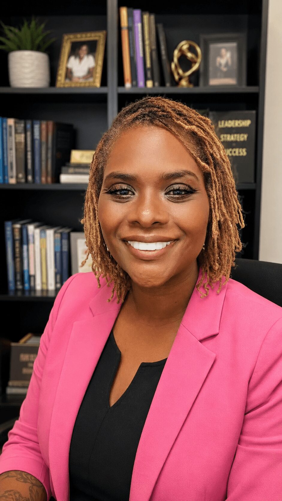 Portrait of a smiling woman with shoulder-length hair, wearing a pink blazer and black top, in an office setting.