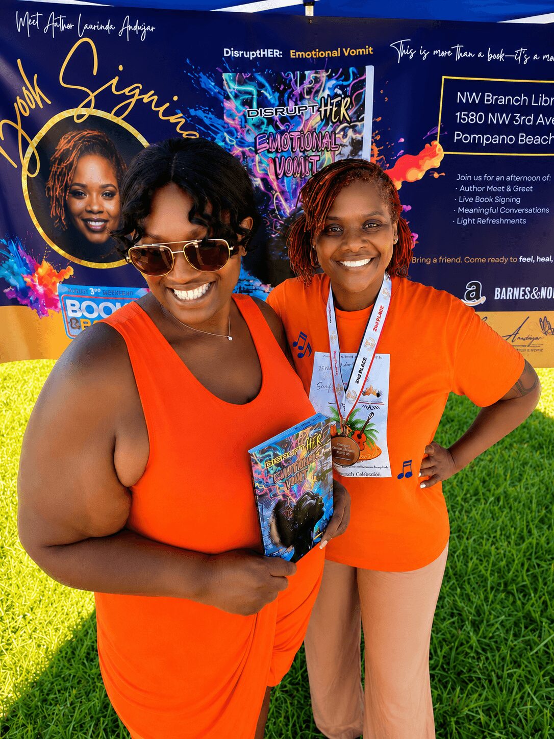 Two women smiling outdoors, one holding a book, both wearing orange tops, with colorful signs in the background.