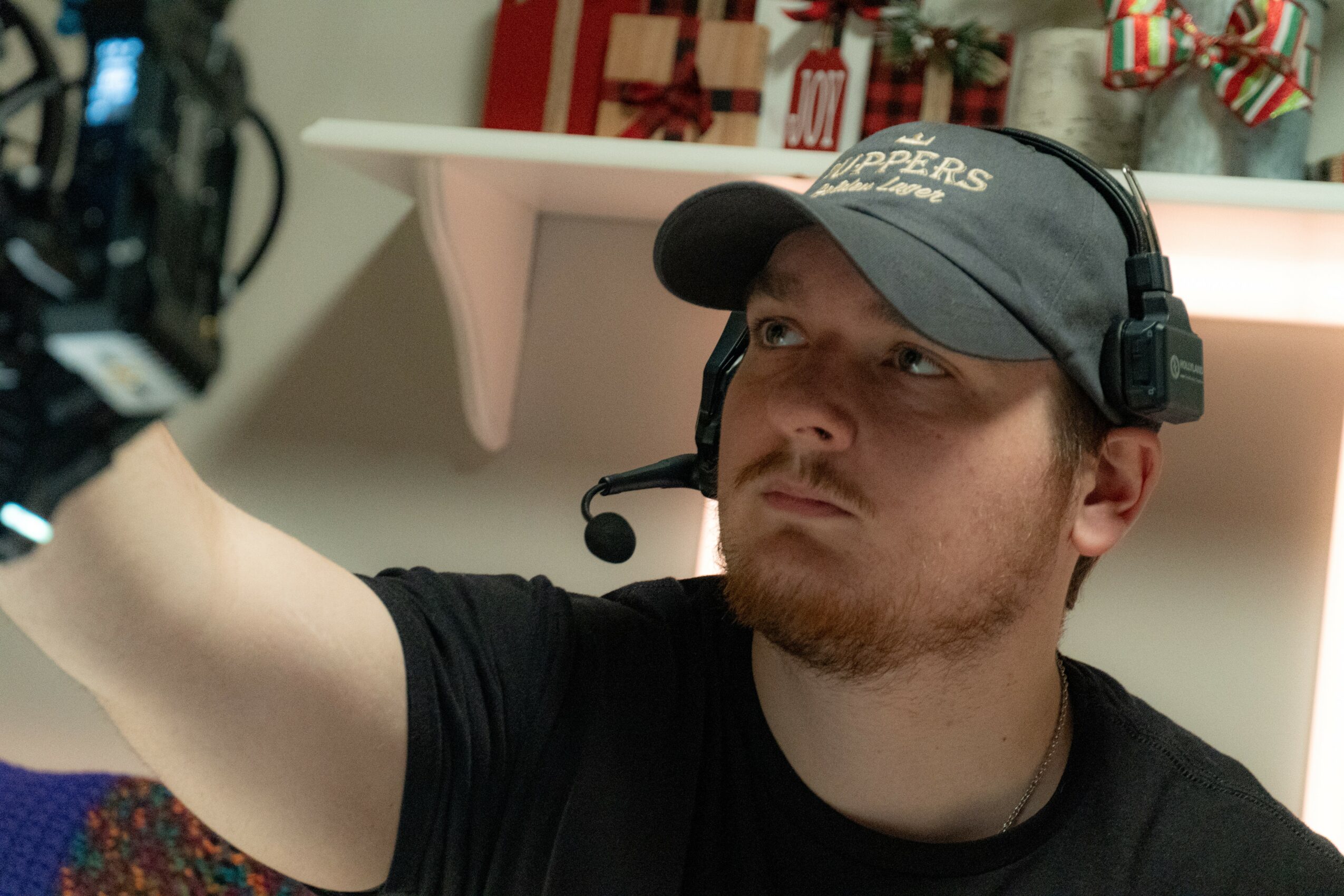 Man wearing a cap and headset, looking to the side, in a room with holiday decorations on a shelf behind him.