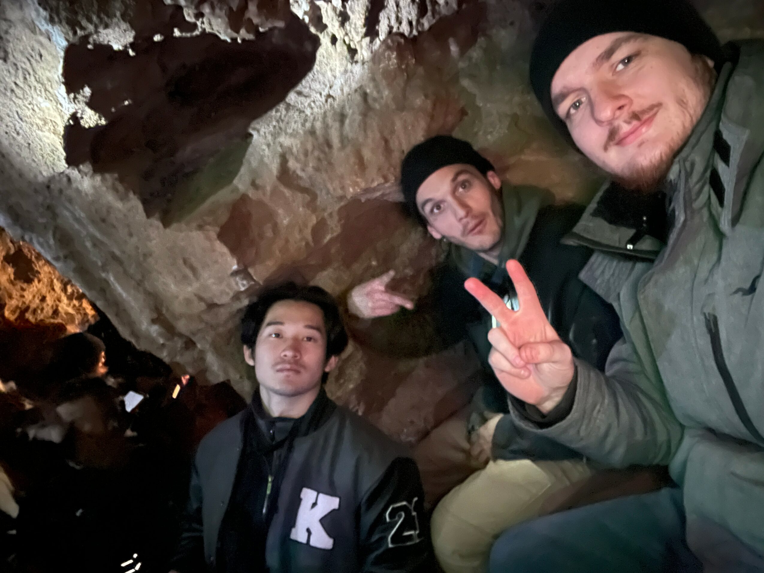 Three people inside a cave, two wearing black beanies, one making a peace sign, all looking at the camera.