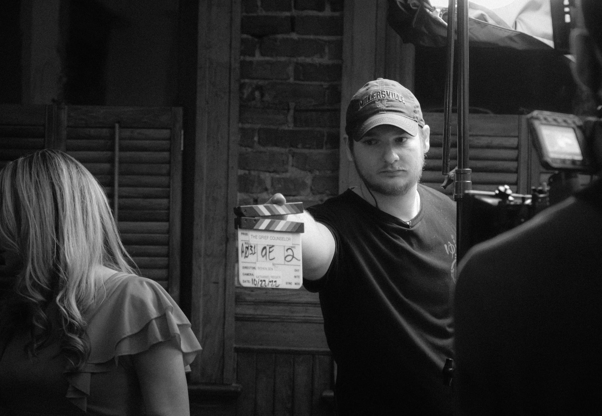 Person holding a clapperboard on set, with a woman in the foreground and a person with a camera in the background.