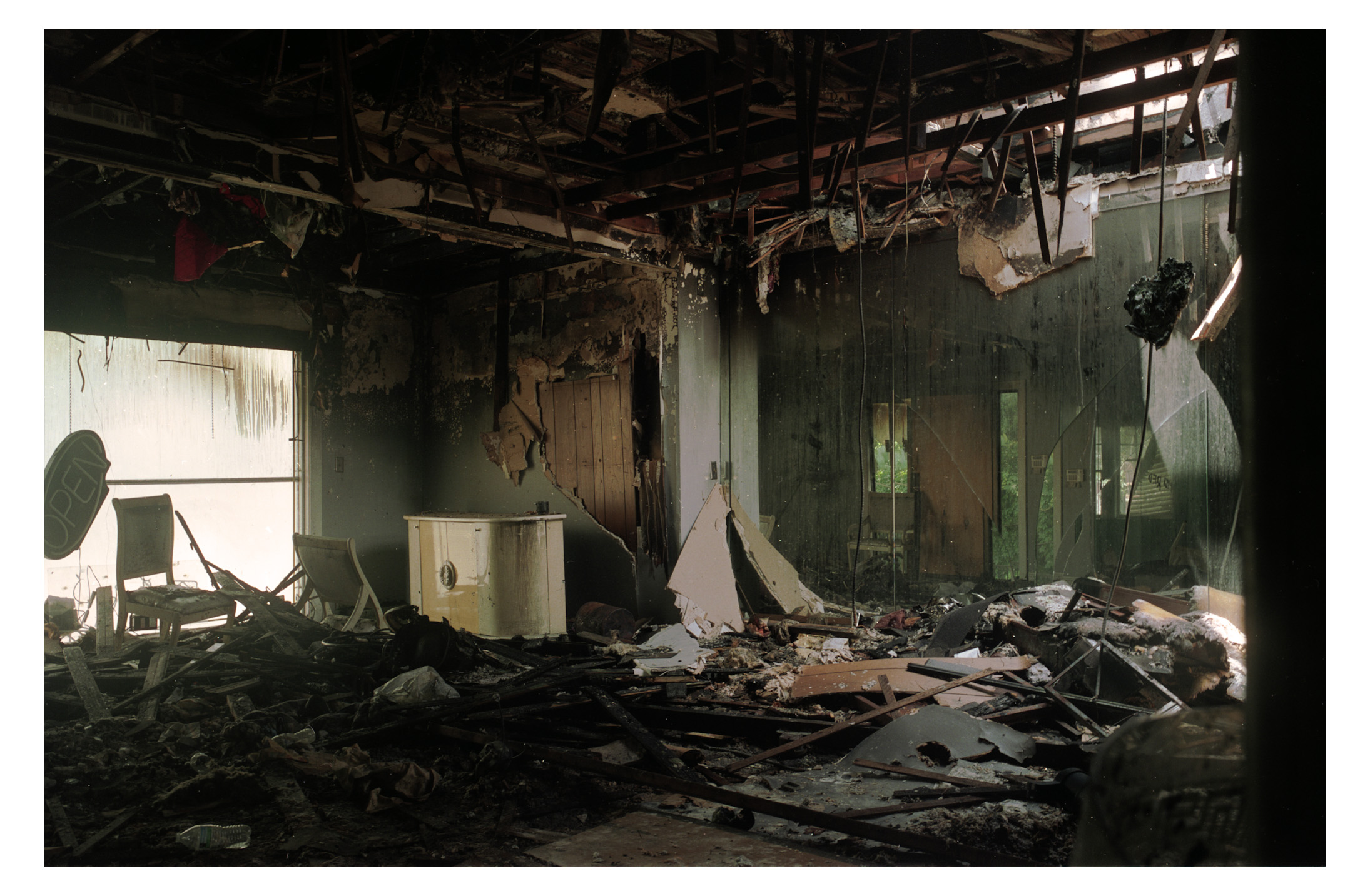 Room with damaged ceiling, debris, and burnt walls, sunlight entering through broken windows, showing destruction inside a building.