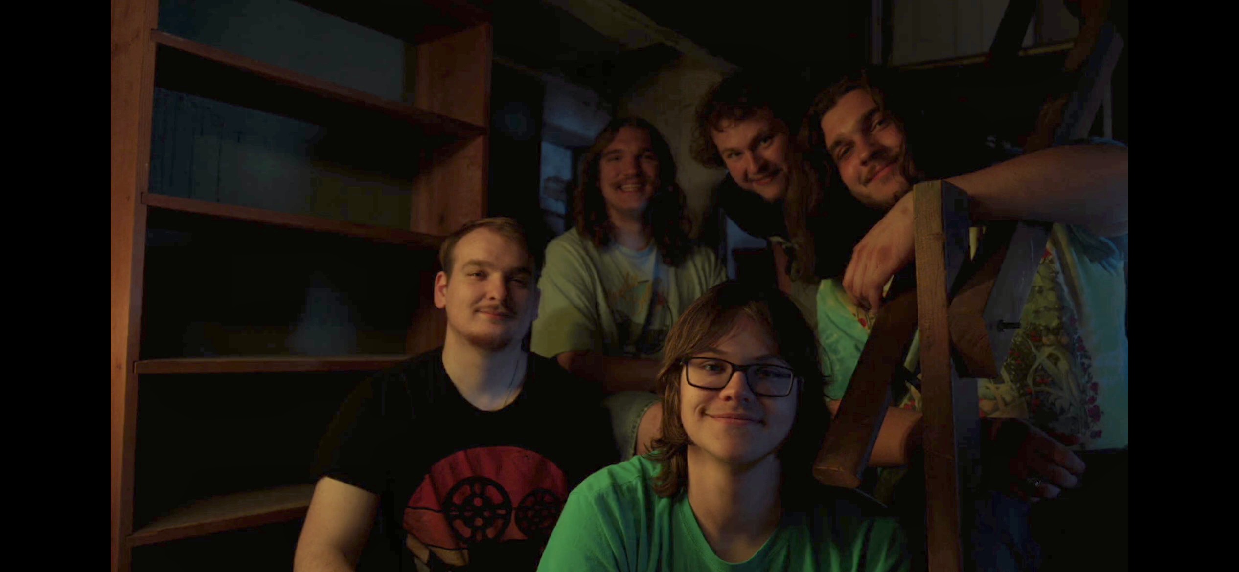 Group of six people gathered indoors, some smiling, in a dimly lit room with shelves in the background.
