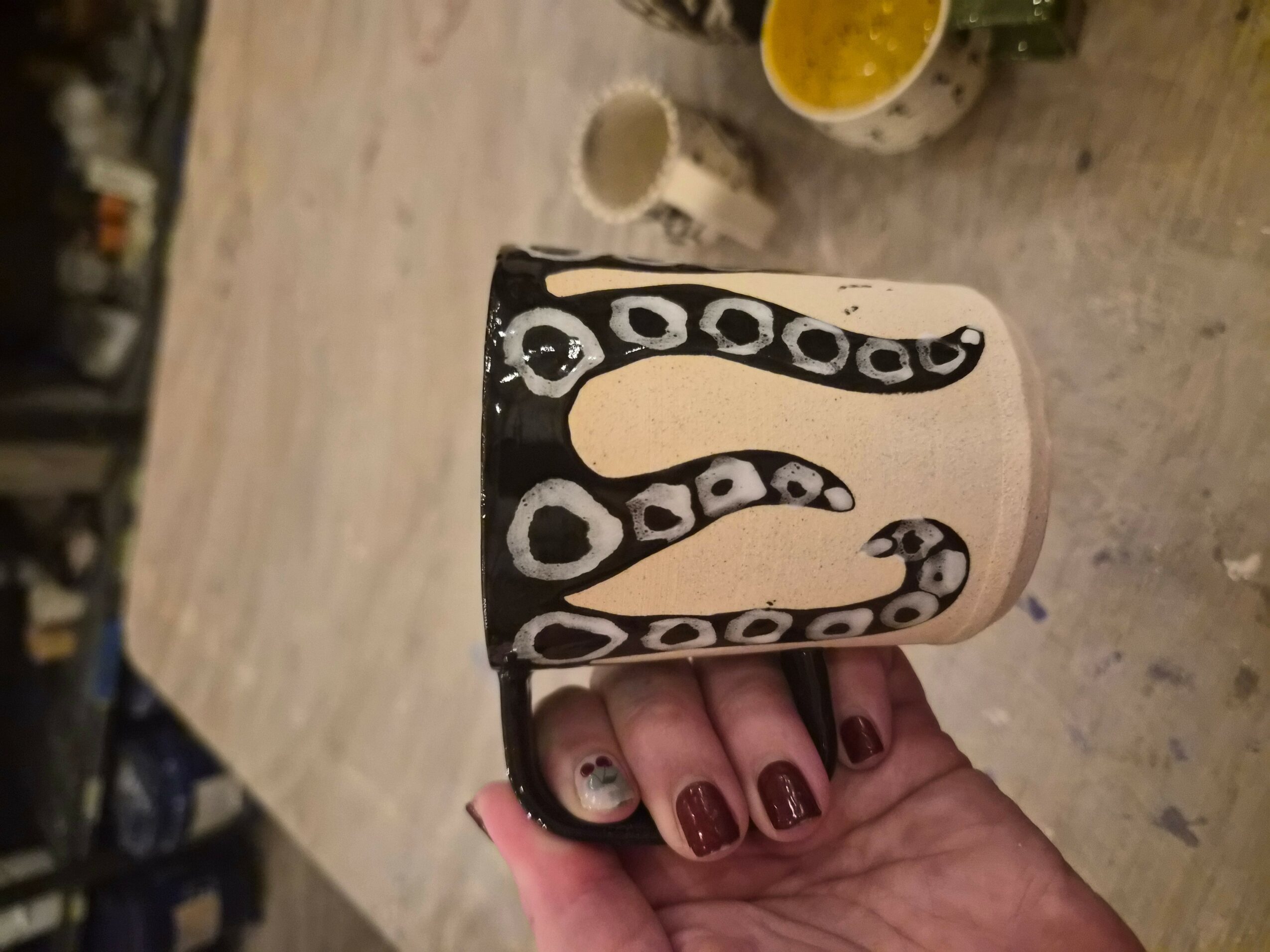 Person holding a mug with a snake design, on a wooden table with cups and a yellow bowl in the background.