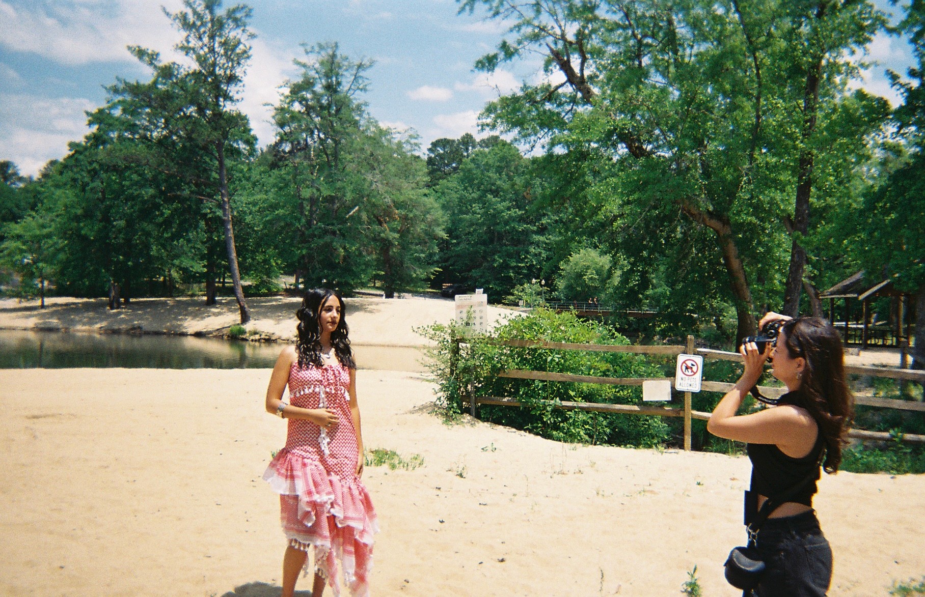 Two women standing outdoors near a body of water with trees and a fence in the background, one taking a photo.
