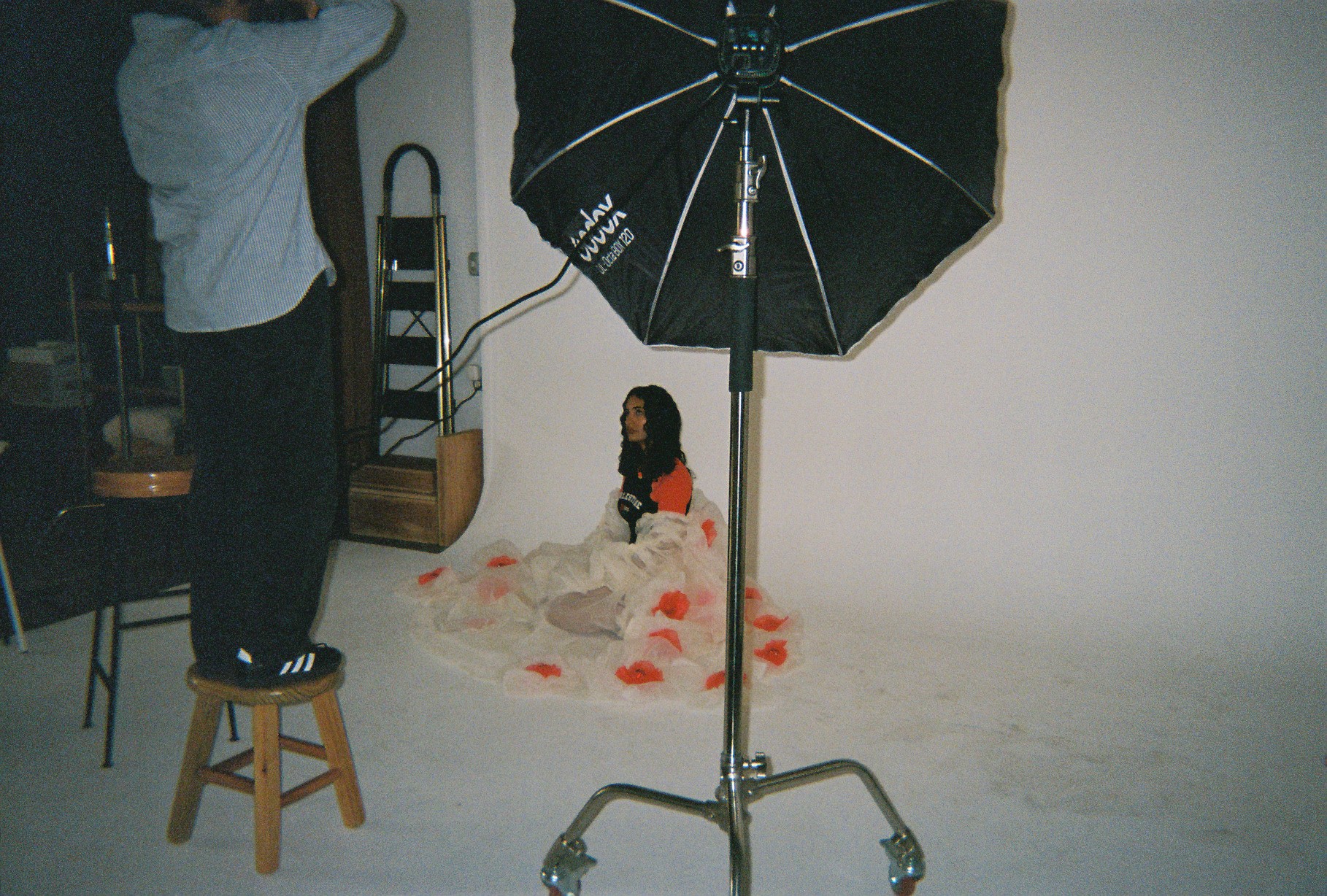 Person standing on a stool adjusting a seated woman in a photography studio with a large umbrella light.