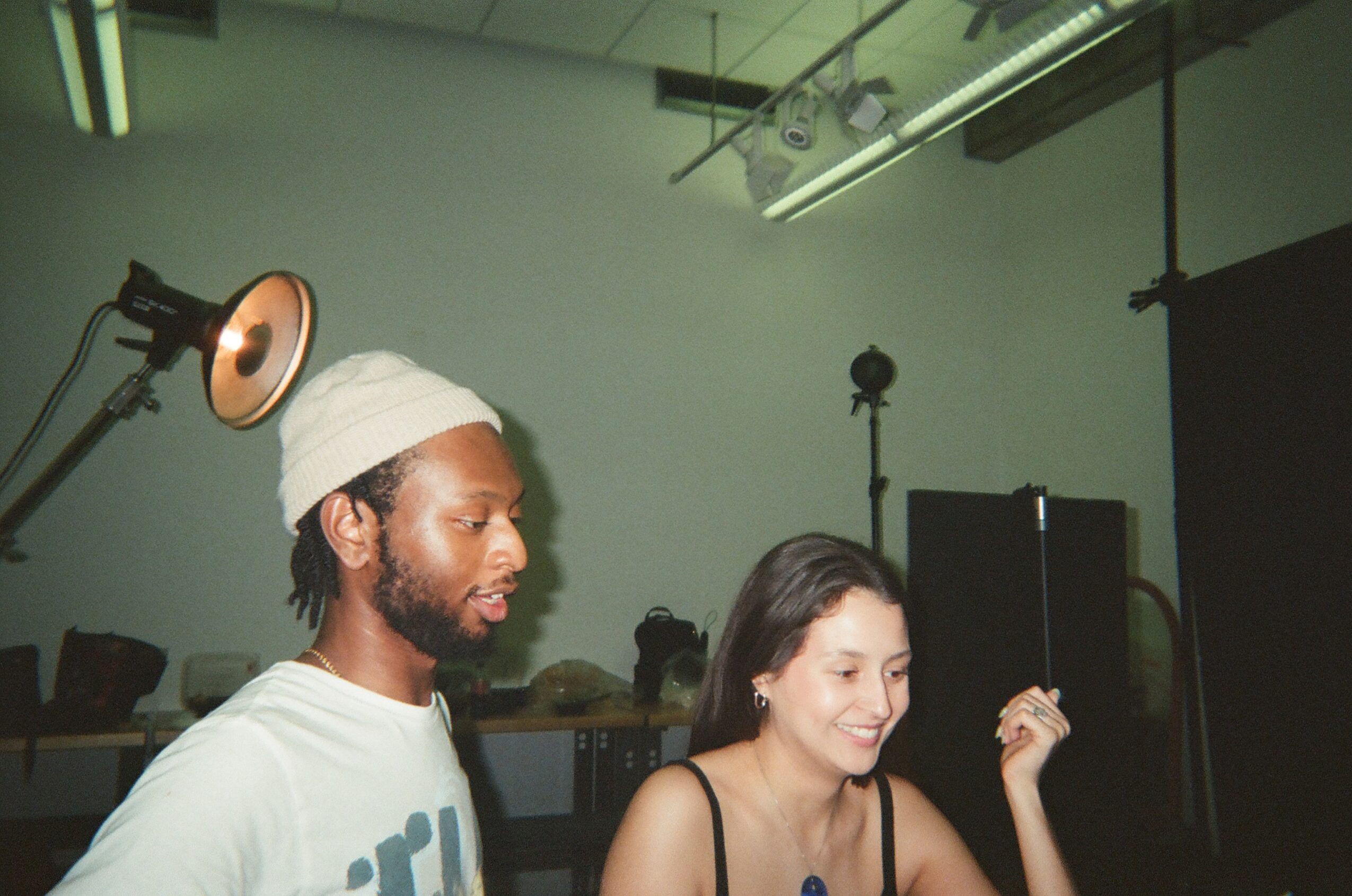 Two people, a man with a white beanie and a woman, are in a studio with lighting equipment and a black backdrop.