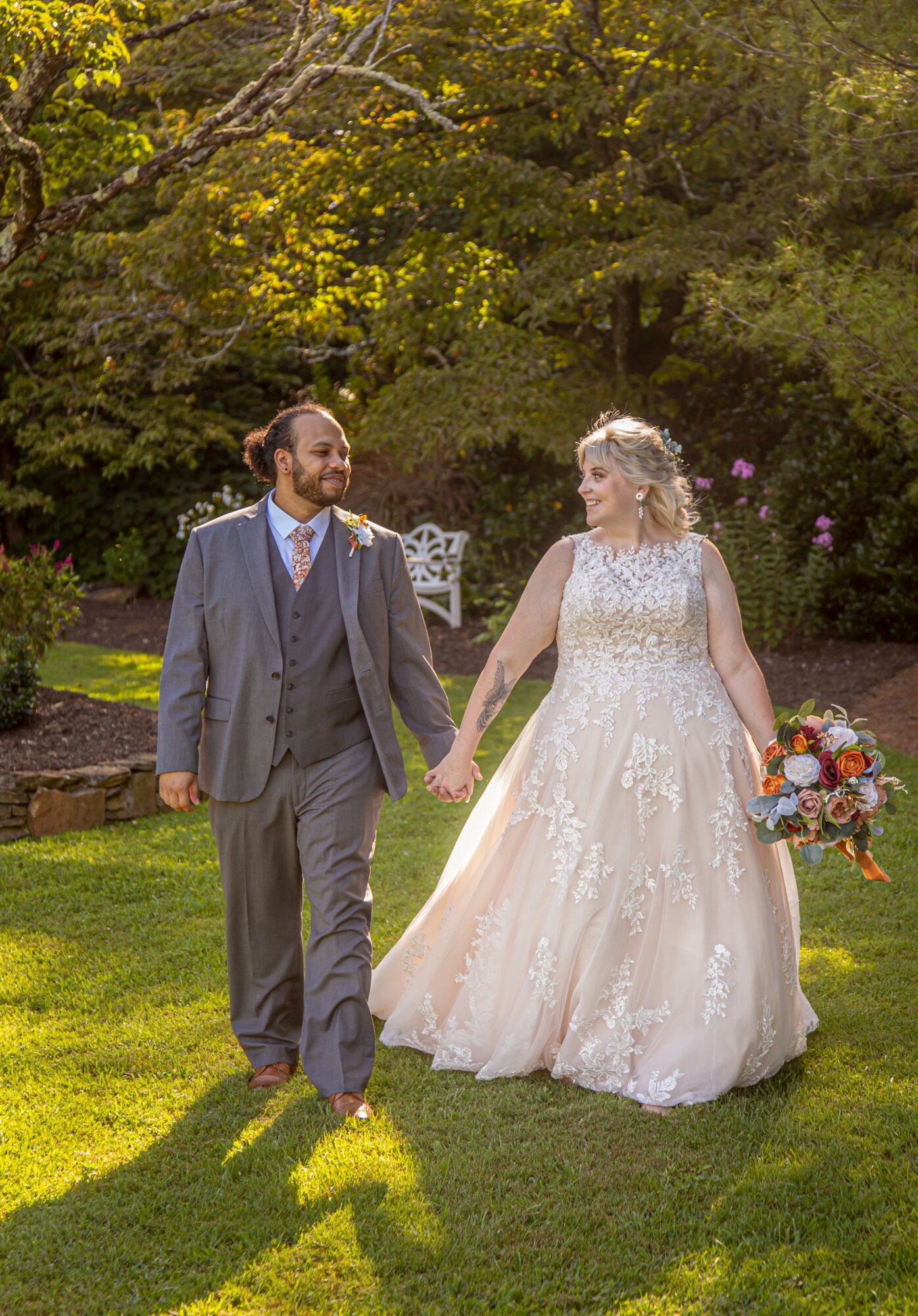 A bride and groom walk hand in hand on a grassy lawn, surrounded by trees, during daytime. The bride holds a colorful bouquet.