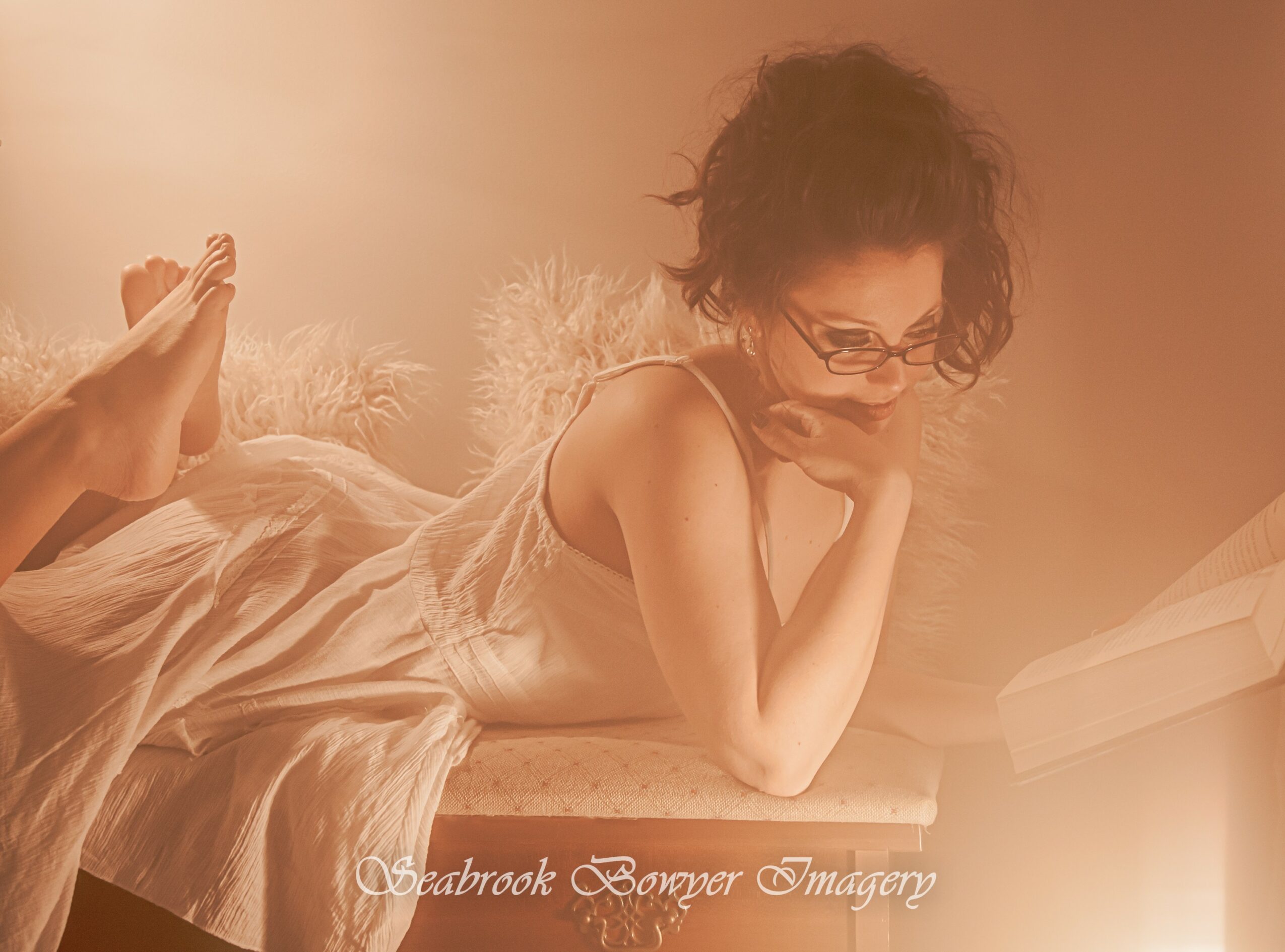 Woman with glasses lying on a bed, resting her head on her hand, in a softly lit room.