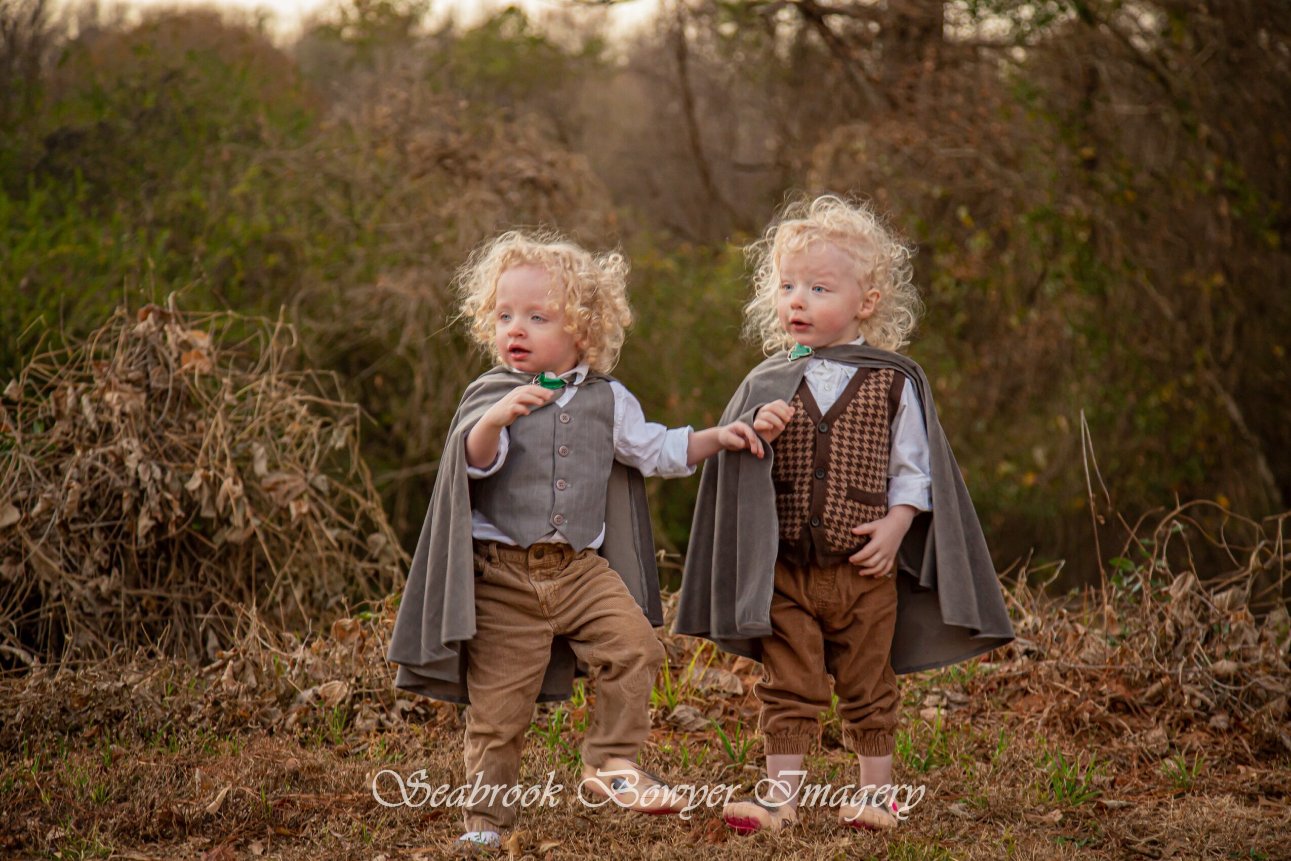 Two children with curly hair wearing capes and vintage-style clothing standing outdoors in a natural setting.