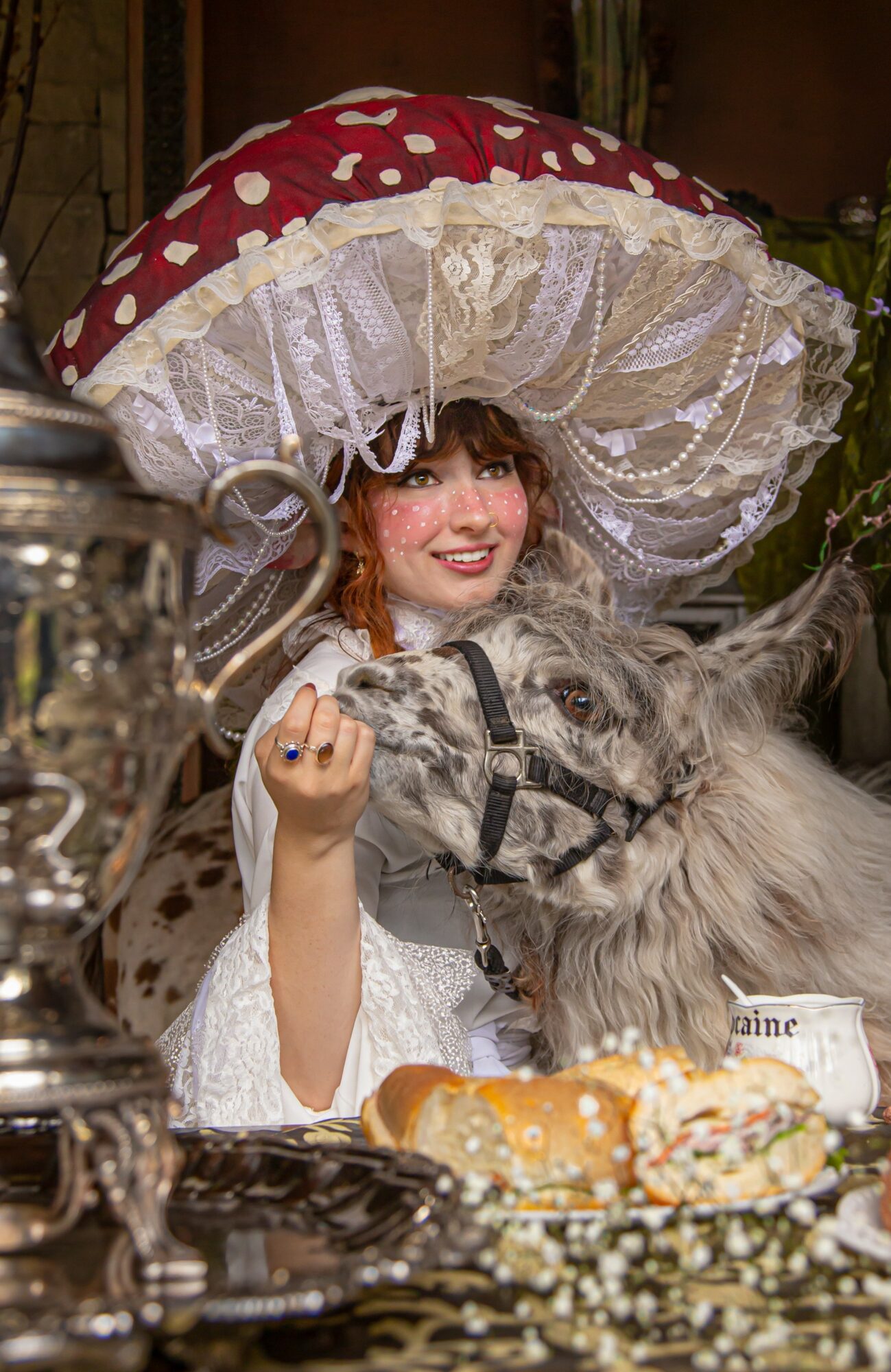 Young woman in a costume with a large mushroom-shaped hat, smiling, holding a small dog, surrounded by food and decorative items.