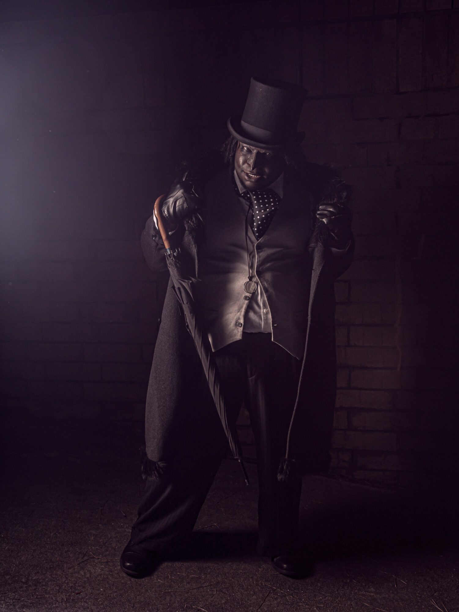 Person dressed in dark clothing with a top hat, holding a cane, standing against a brick wall, dimly lit.
