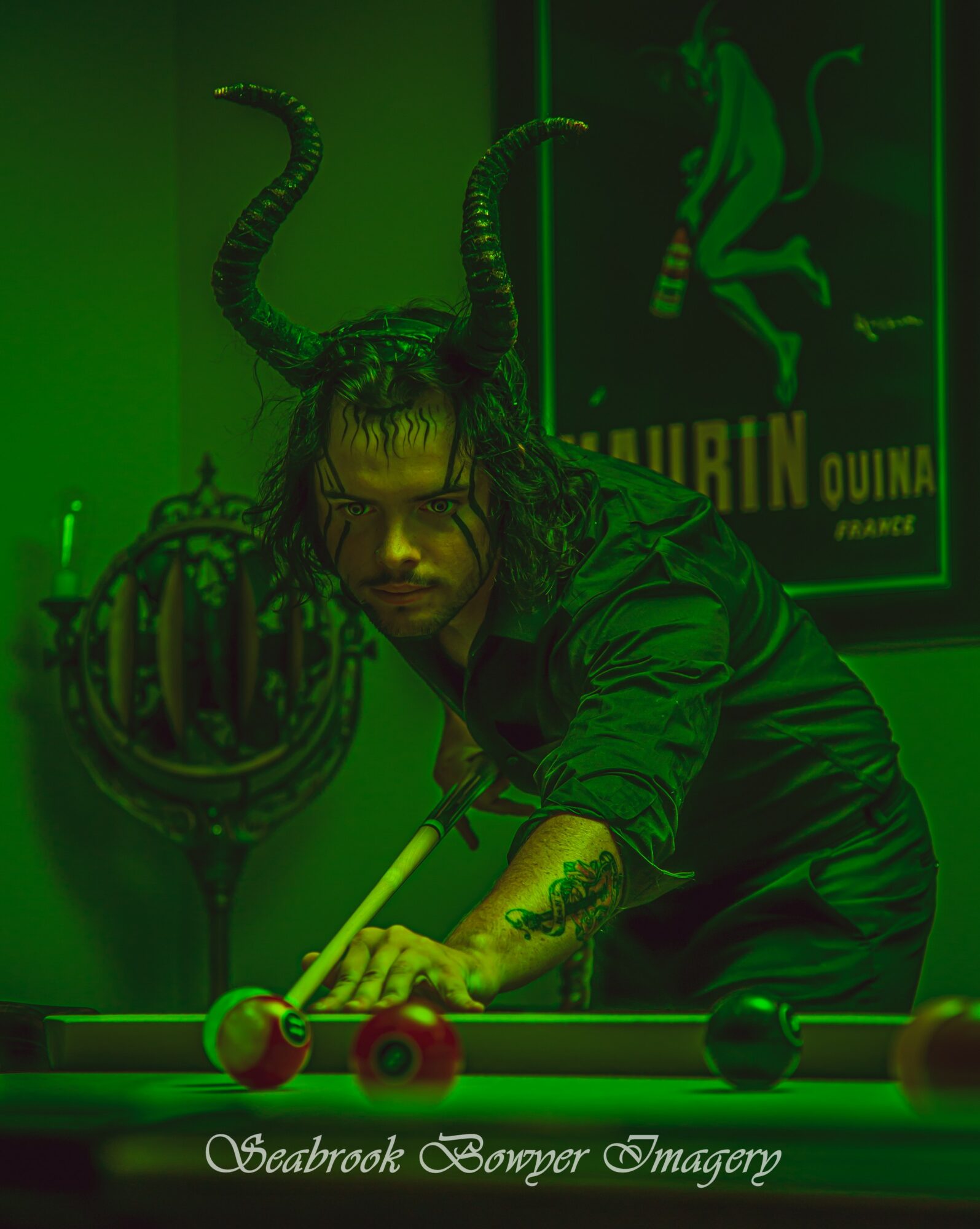 Person with long hair and tattoos playing pool, with a poster and a sculpture of a creature with horns in the background.