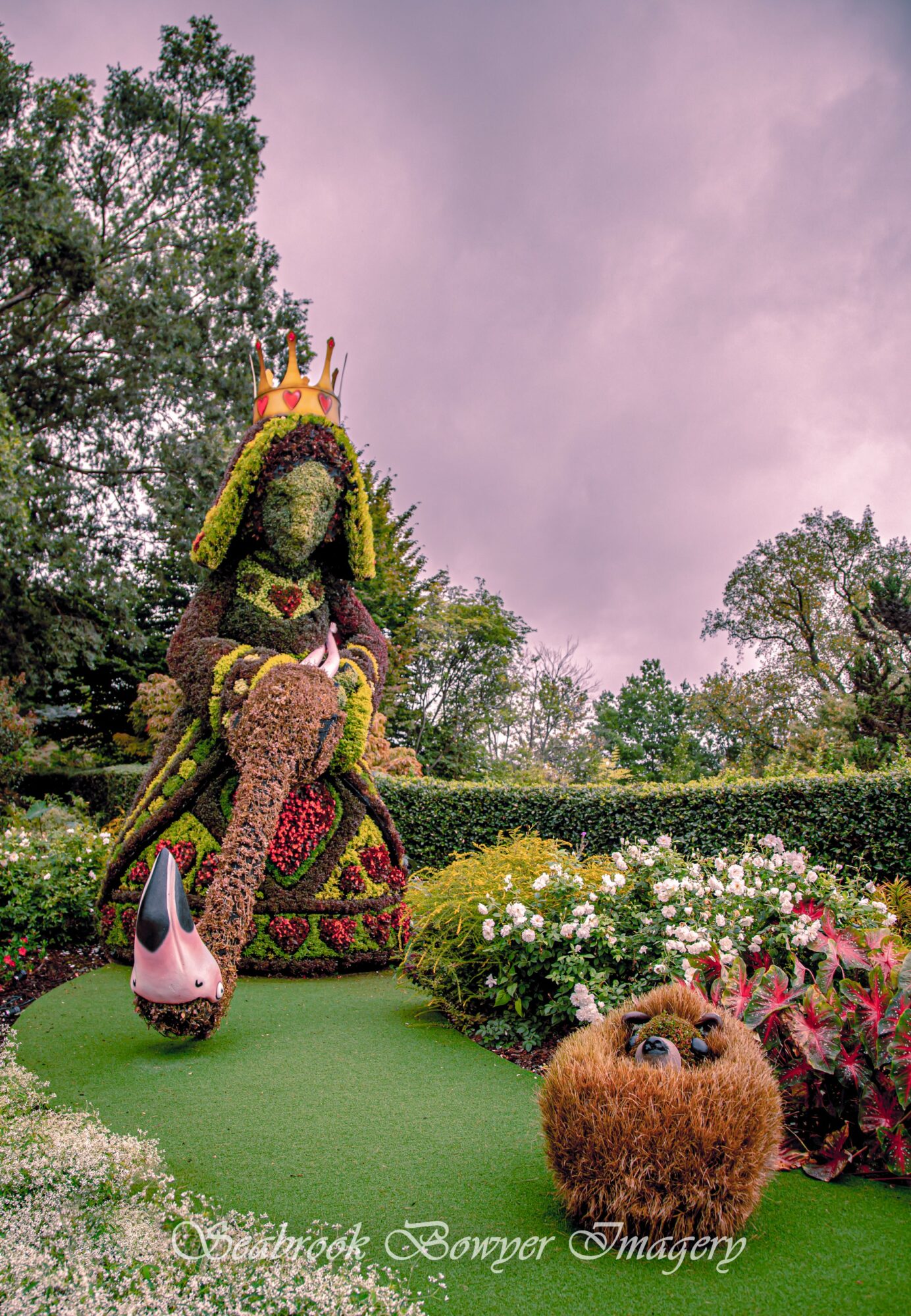 Decorative floral sculpture of a woman with a crown, surrounded by flowers and a small dog figure, outdoors with trees and sky.