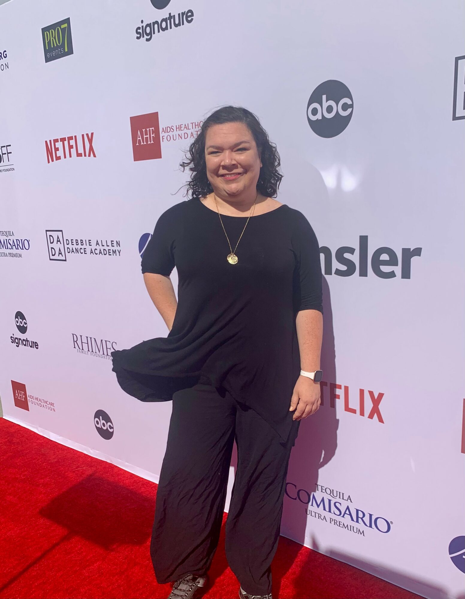 Smiling woman in black outfit standing on red carpet with event backdrop behind her.