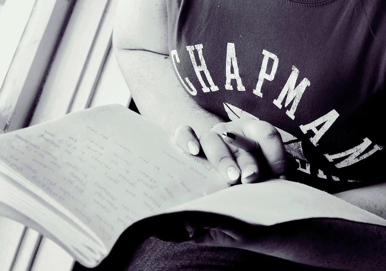 Person reading a book with hands clasped, wearing a shirt with partially visible text, in black and white.