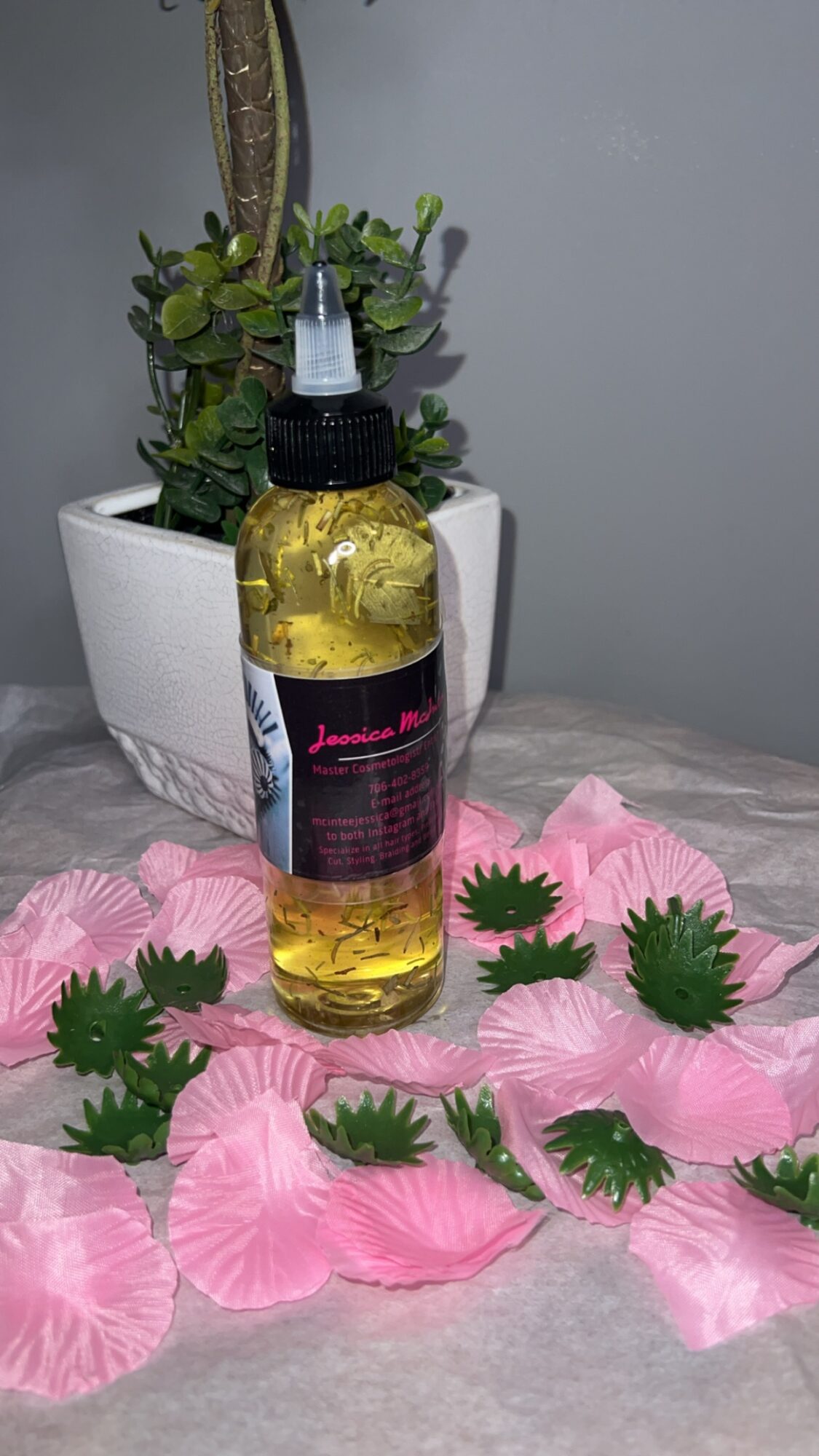 A small bottle with a dropper cap placed on pink flower petals and green leaves, with a potted plant in the background.