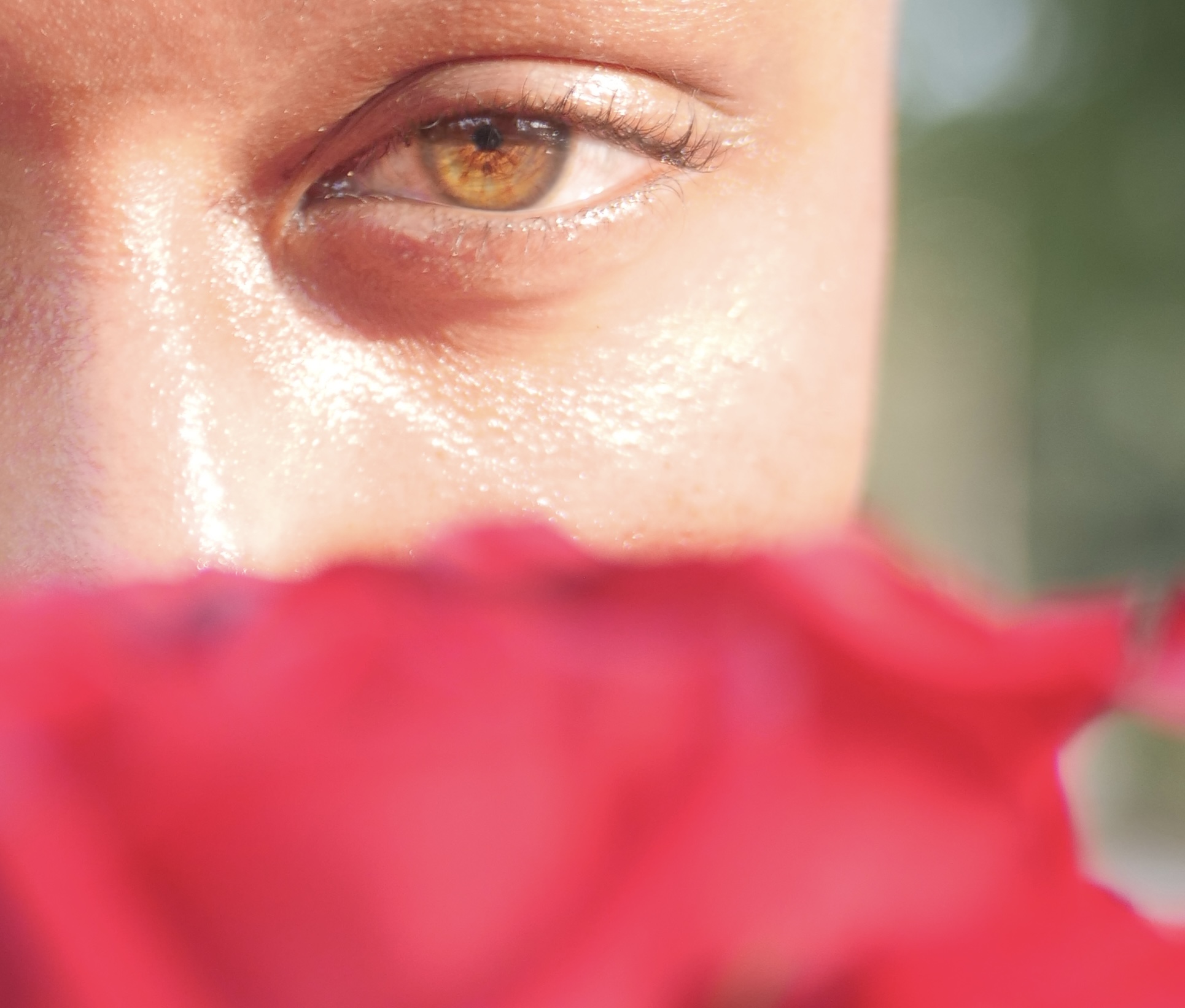 Close-up of a person's eye with a blurred red flower in the foreground, outdoors with natural light.