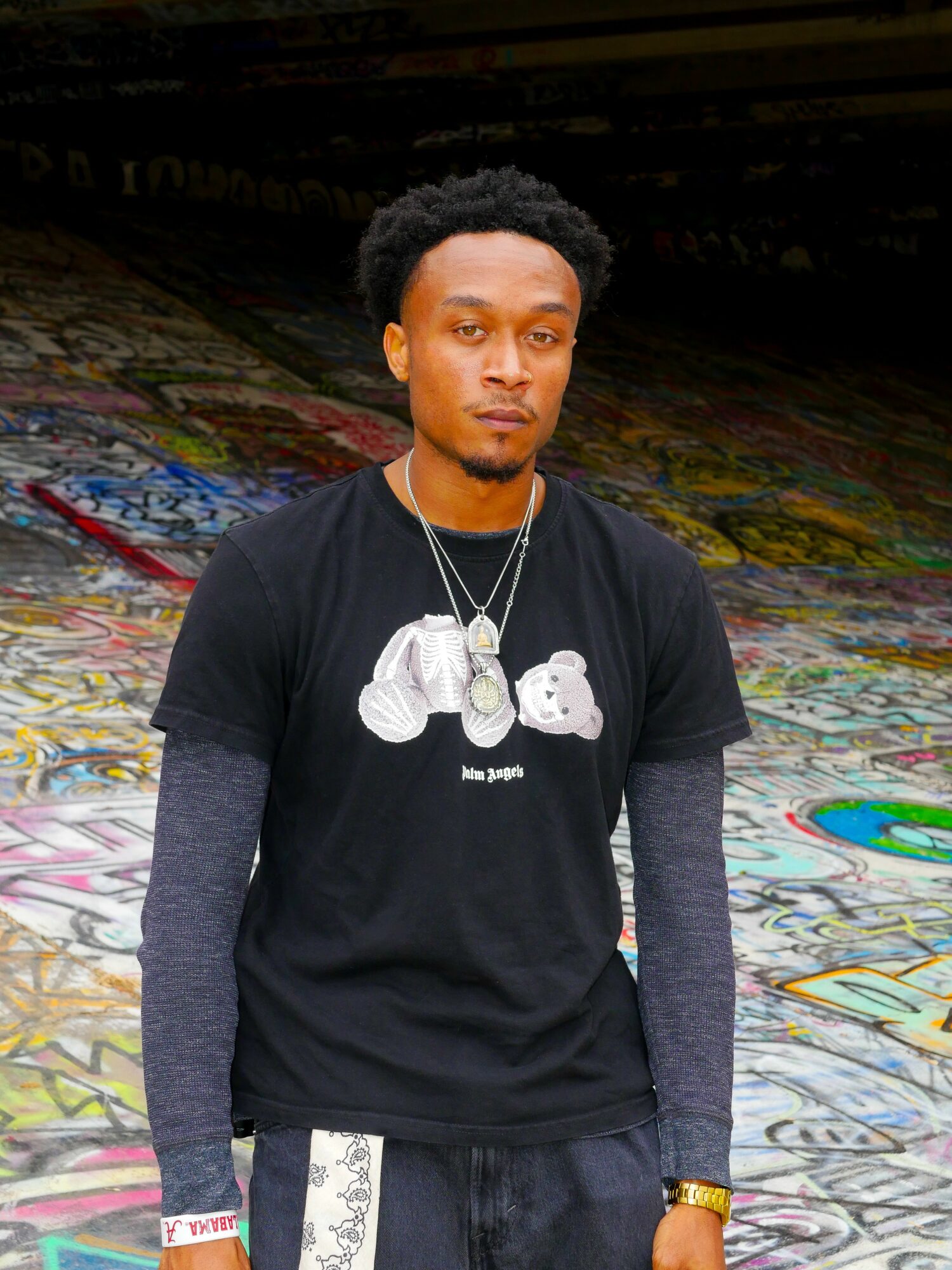 Young man with short curly hair stands on graffiti-covered ground, wearing a black t-shirt with cartoon characters, layered gray sleeves, and jewelry.