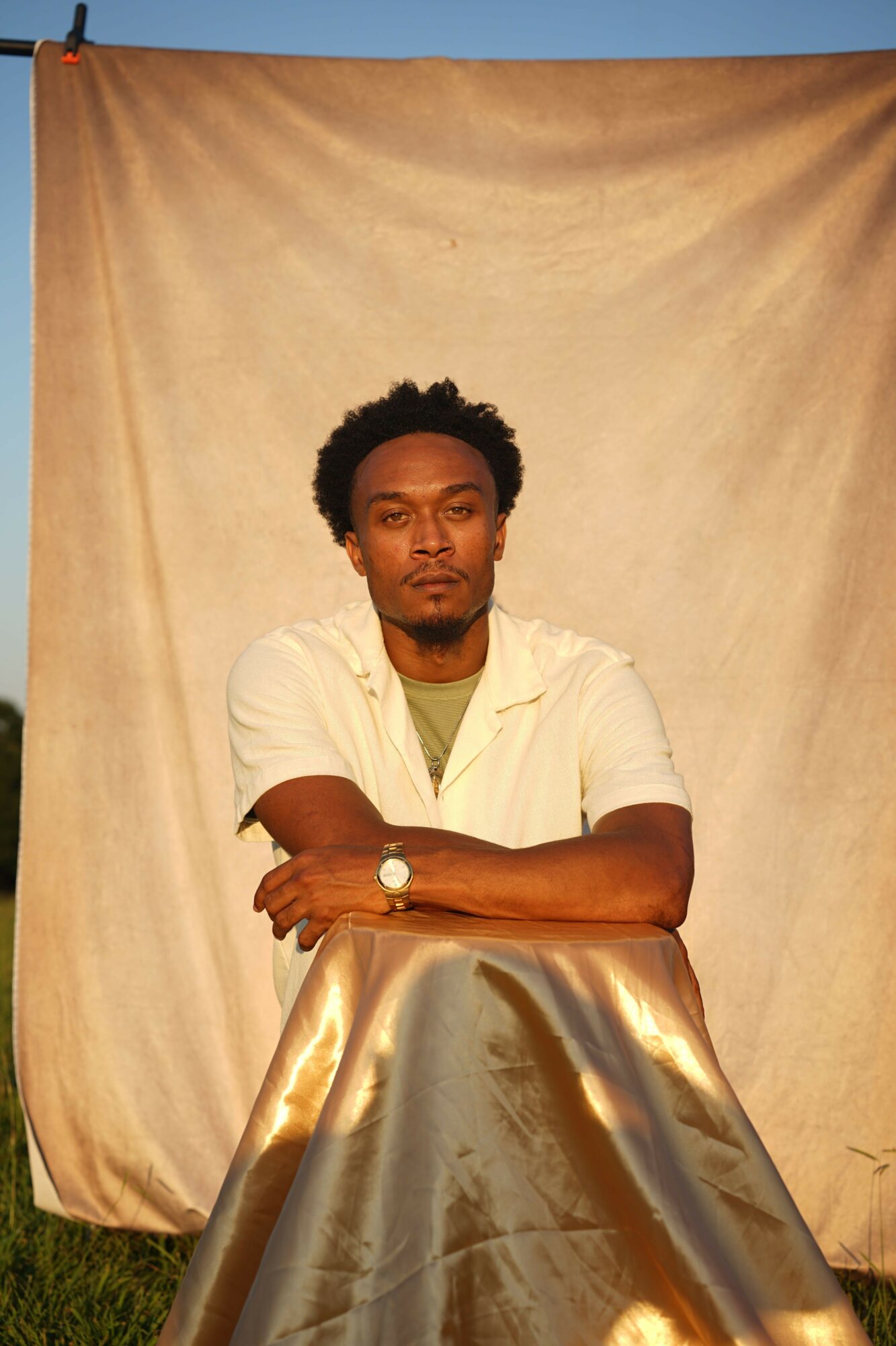 Person with short curly hair leaning on a reflective surface, wearing a white shirt, against a beige backdrop.