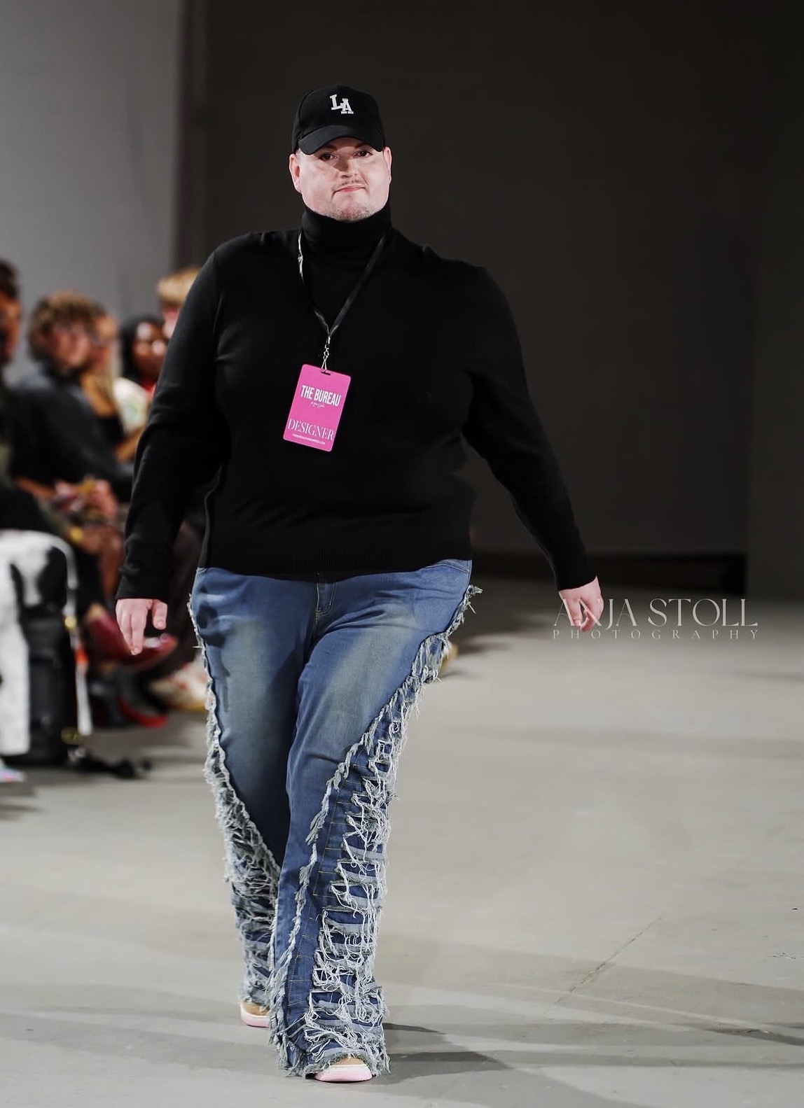 Person walking on runway wearing black top, jeans with lace-up design, black cap, and pink badge, audience seated in background.
