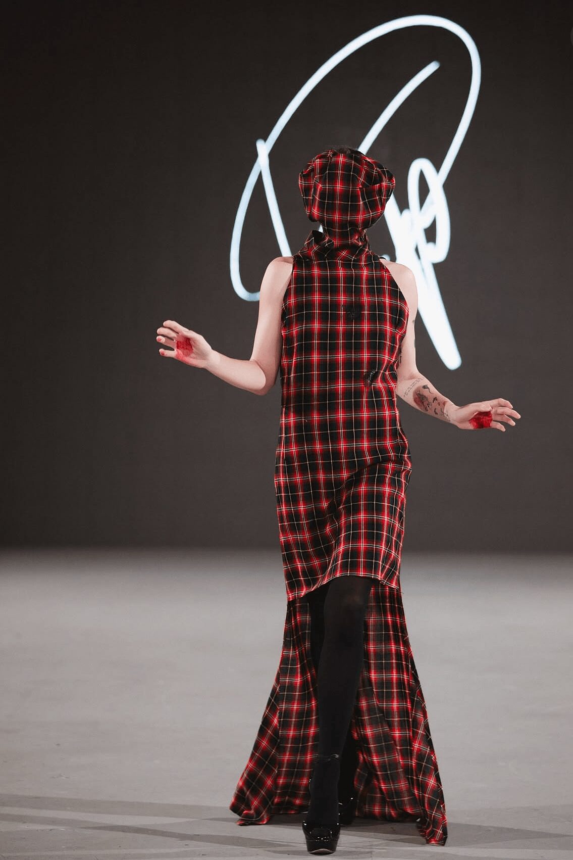 Person walking away in a long plaid dress and matching hat, with a dark background and white abstract design. Word count: 22