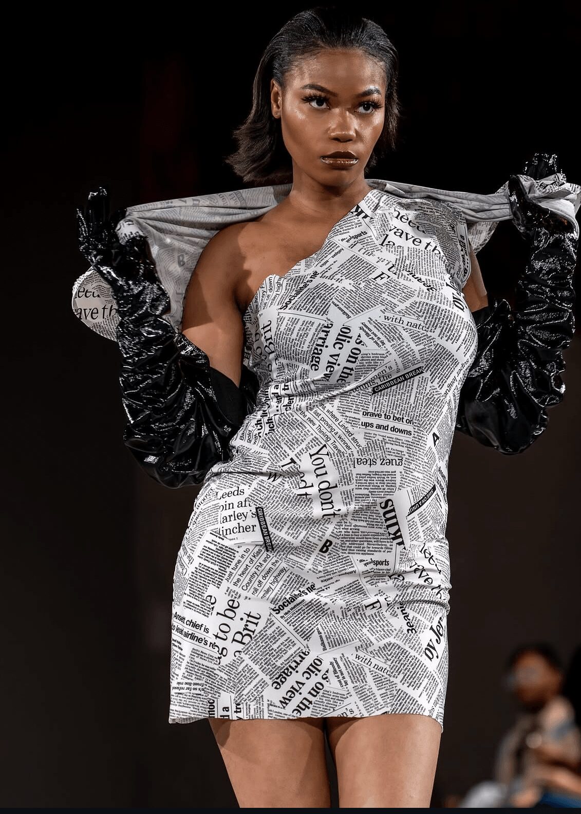 Model wearing a newspaper print dress and black gloves, holding a transparent umbrella, walking on runway.