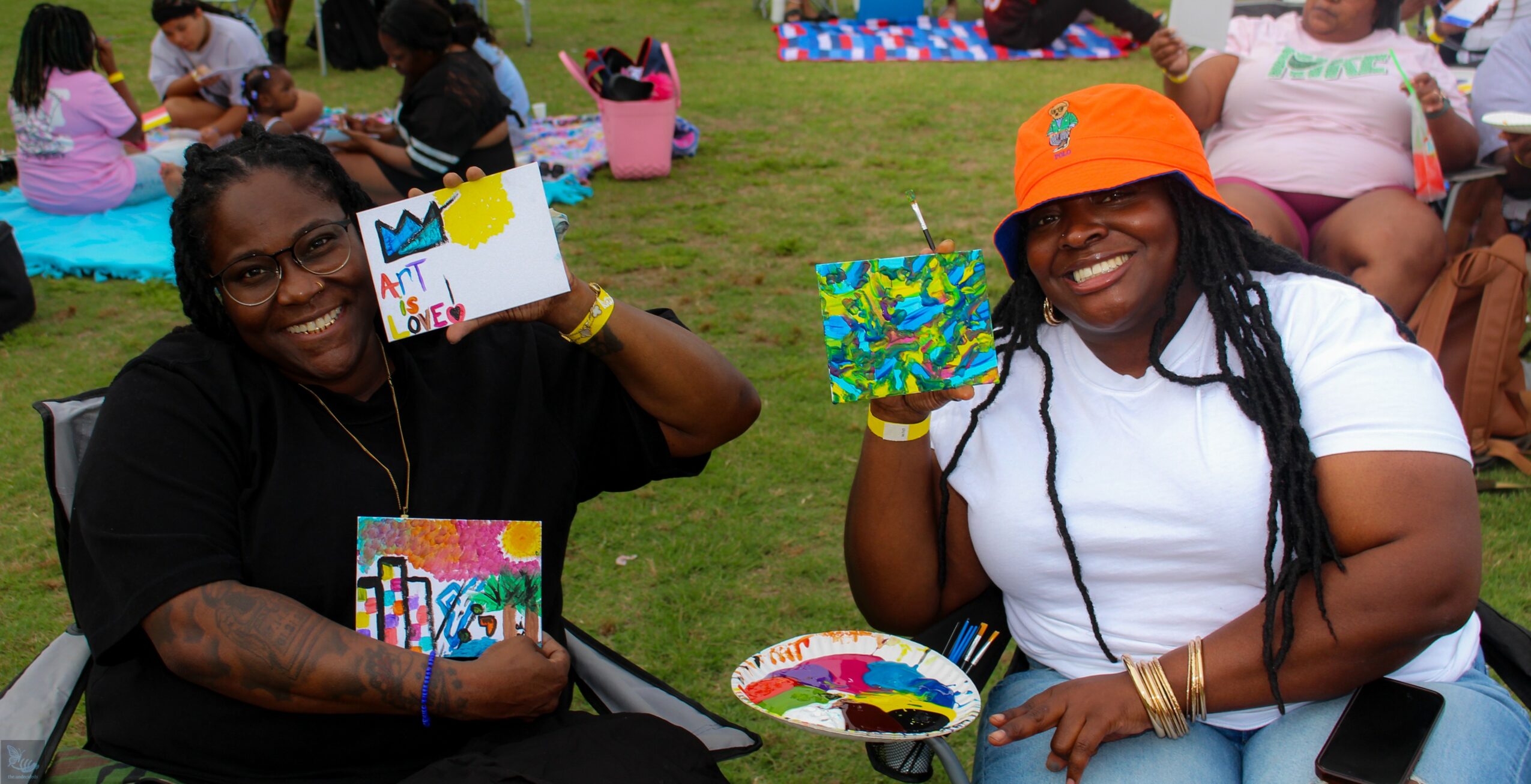 Two women sitting outdoors holding colorful painted canvases, smiling, with other people in the background, on a grassy area.