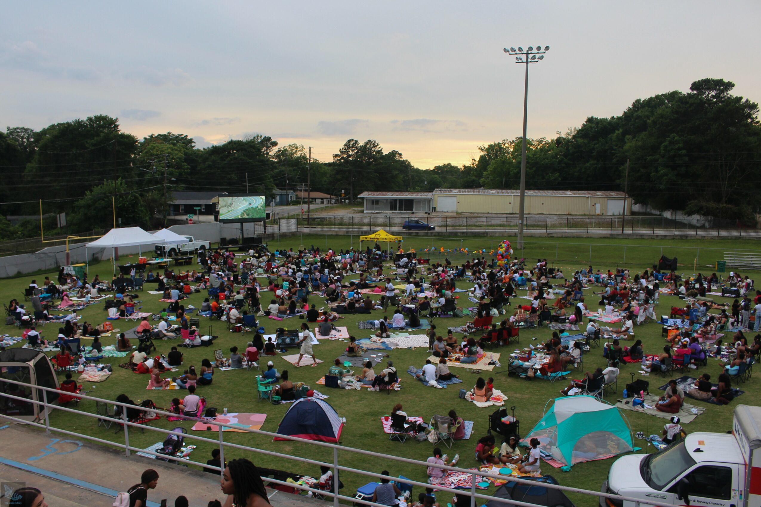 Large outdoor gathering with many people sitting on grass, some under umbrellas, in a park or sports field.