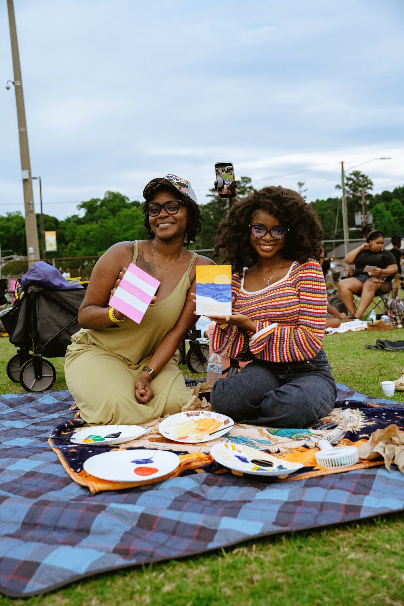 Two women sitting on a blanket outdoors, holding colorful paintings, with people and trees in the background.