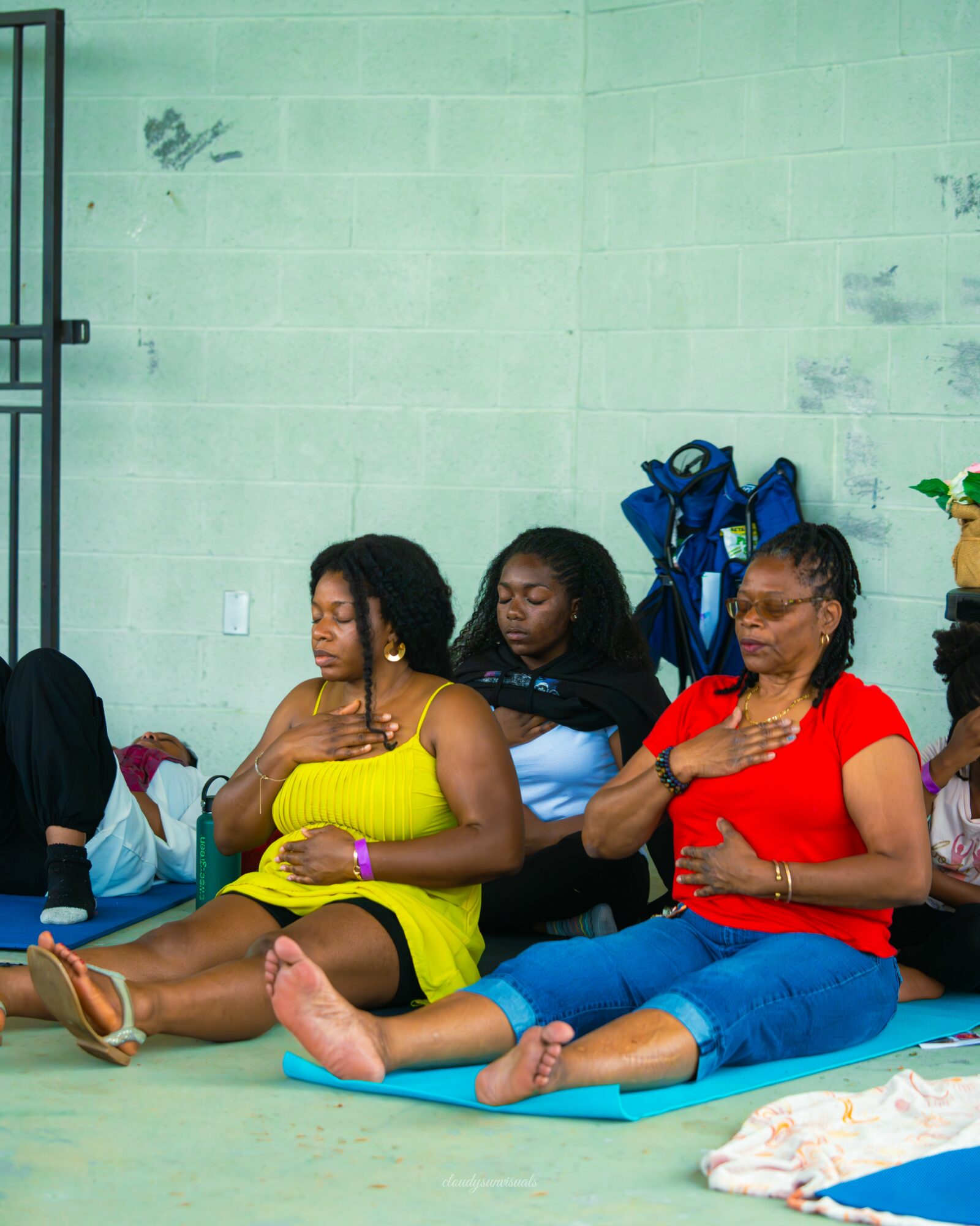 Three women sitting cross-legged on a mat, practicing meditation or mindfulness with hands over their hearts, against a light green wall.