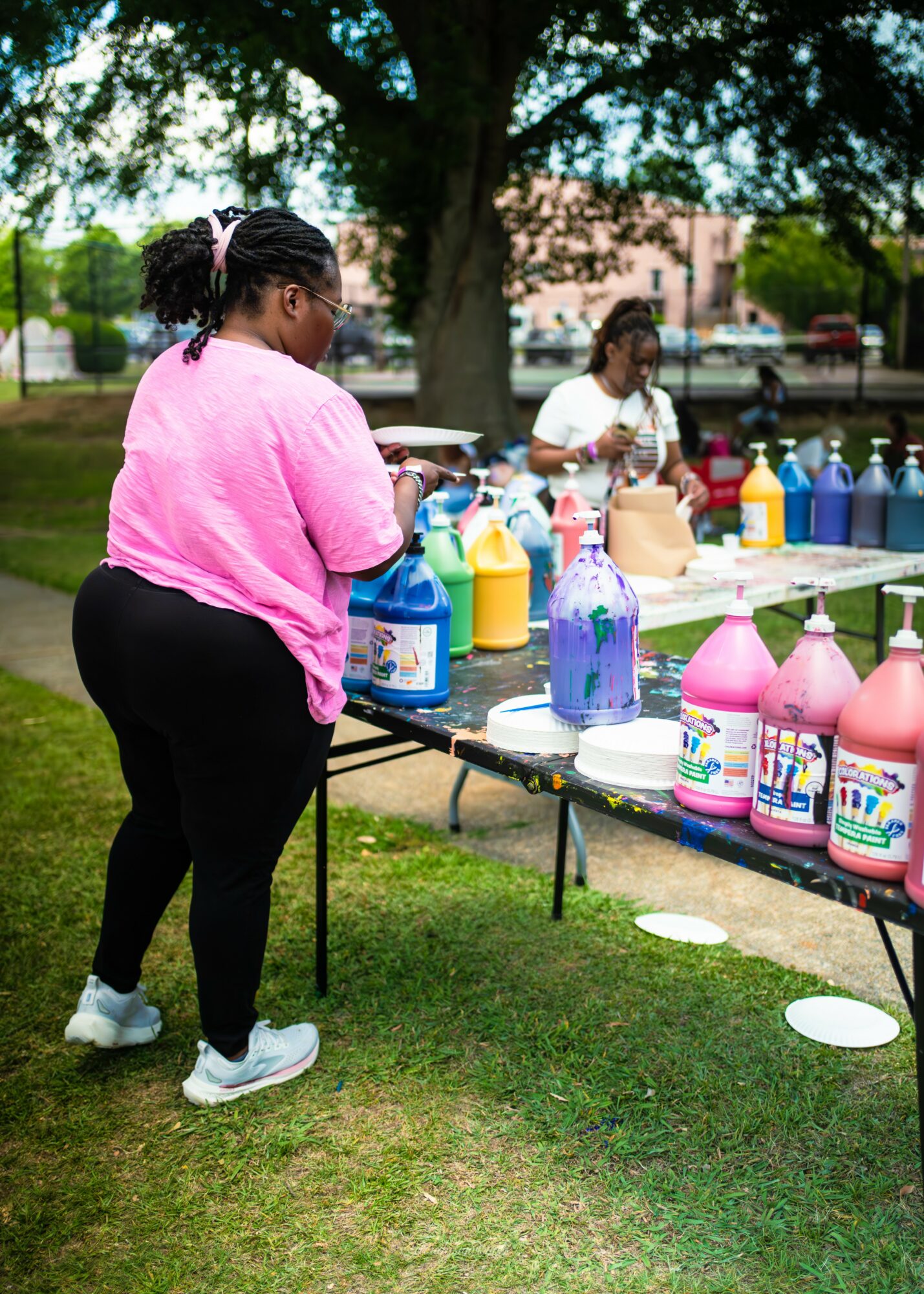 Two women at an outdoor table with colorful bottles, trees and buildings in the background, one woman in pink facing the table.
