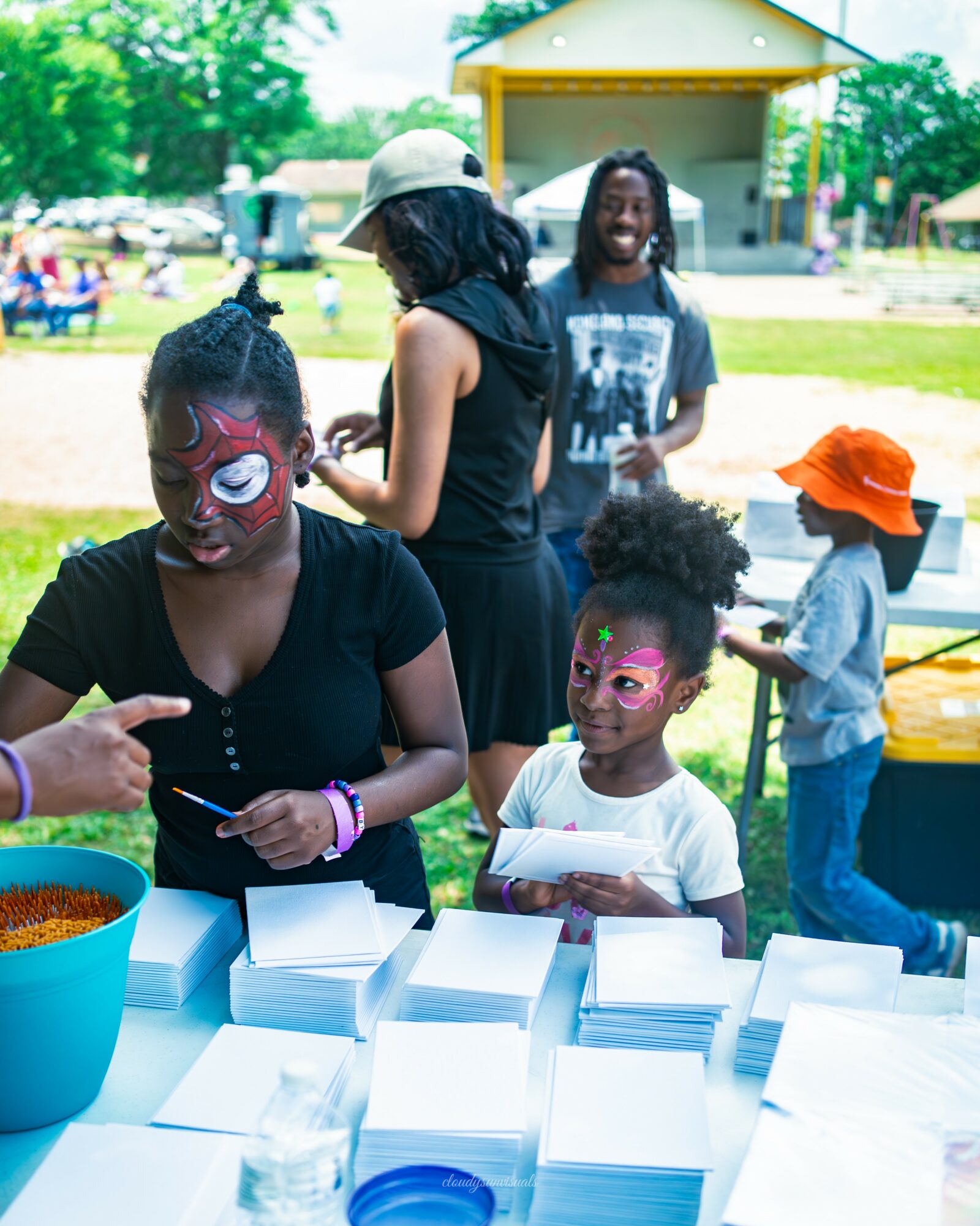 Children and adults outdoors at a park with face paint, tables, and a yellow building in background.