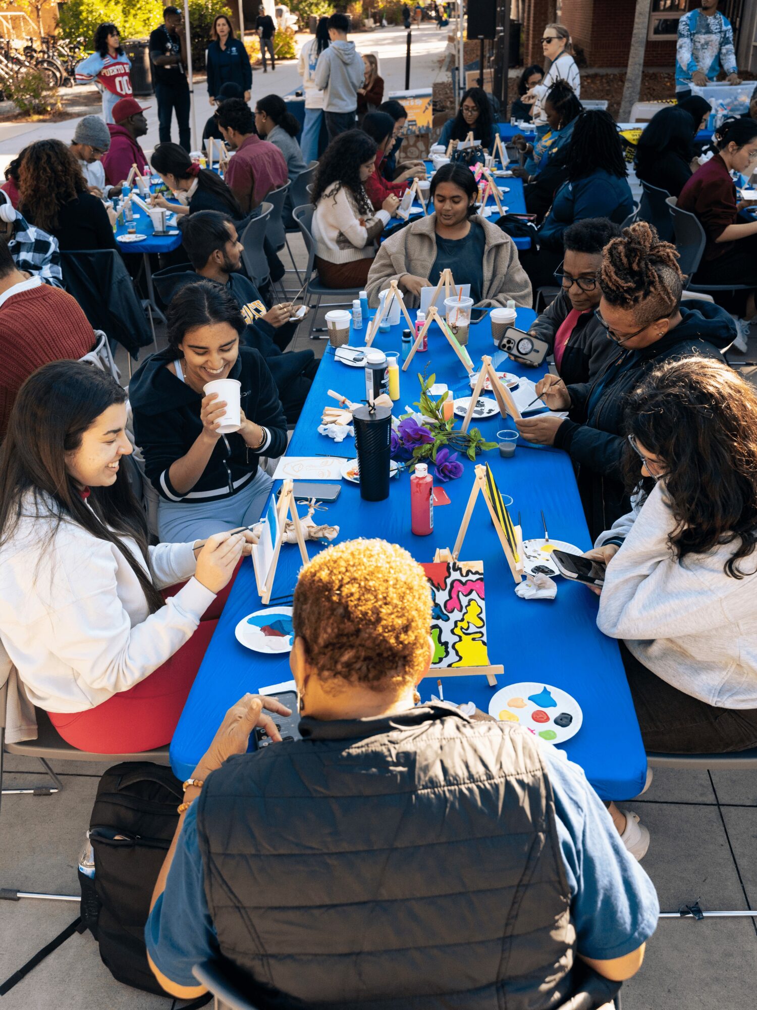 People gathered around a long table outdoors, engaging in arts and crafts activities, with some standing and others seated.