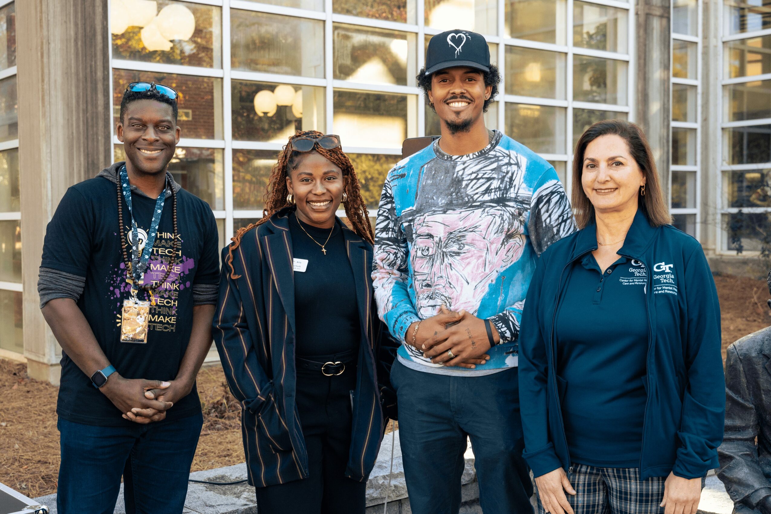Four diverse people standing outdoors in front of a building with large windows, smiling and posing for the photo.