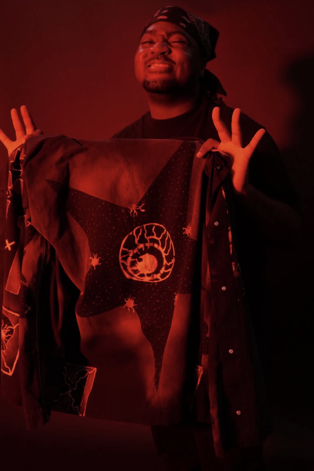 Person smiling with hands raised, wearing a dark outfit and a head covering, illuminated by red lighting.