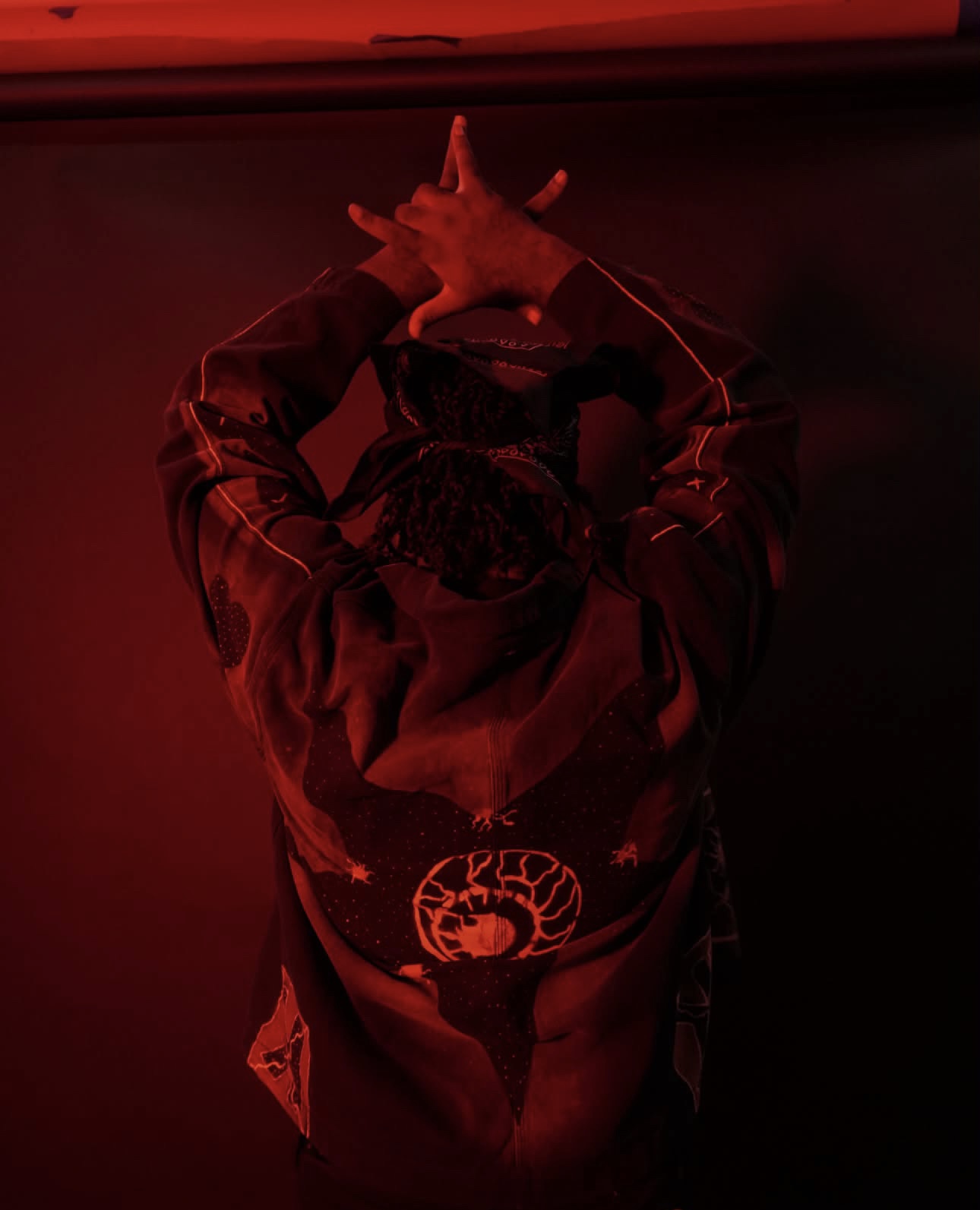 Person wearing a hoodie with a graphic design, standing in a dark red-lit environment, hands on head, facing away.