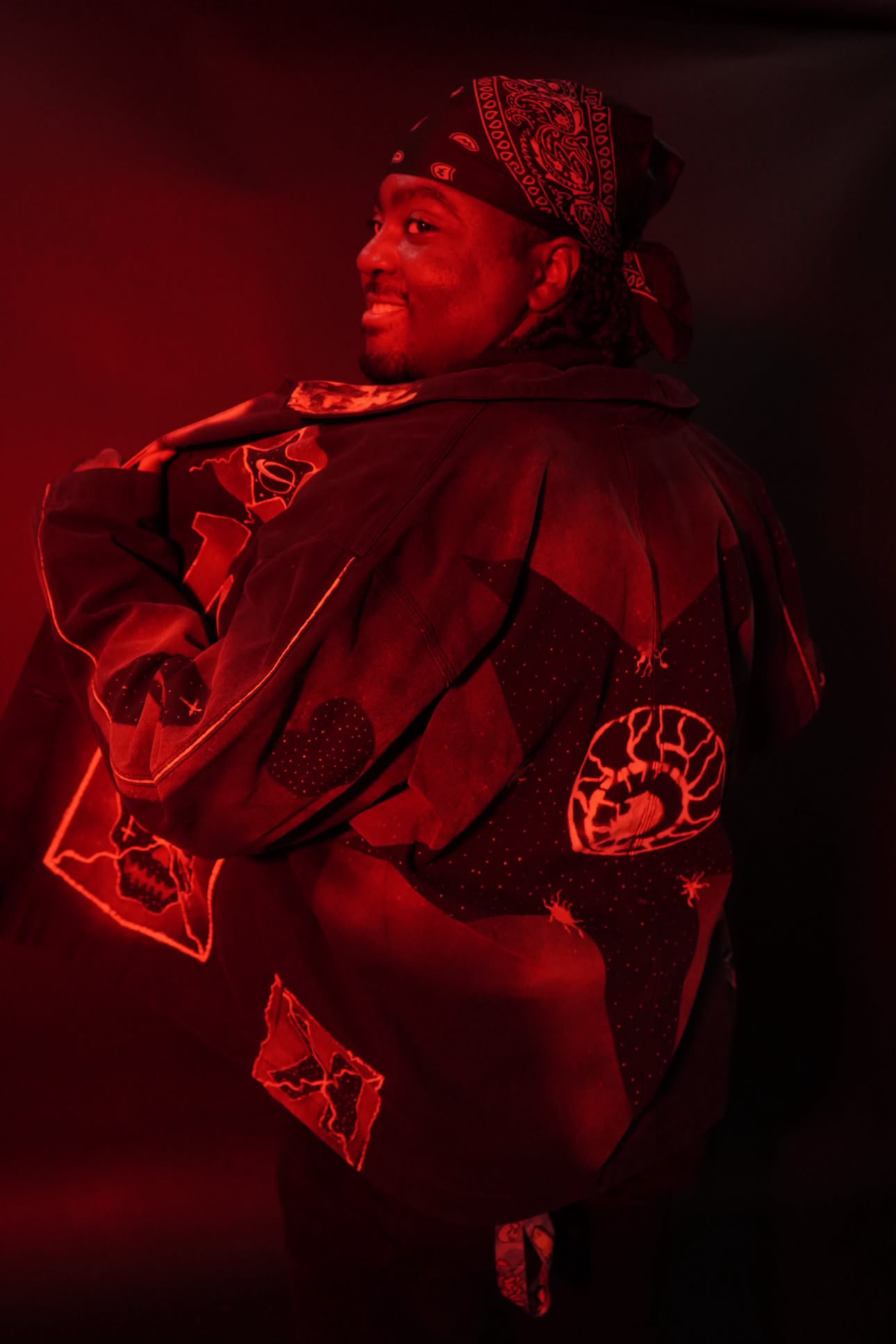 Person smiling, wearing a bandana and jacket with patches, illuminated by red light, facing sideways.