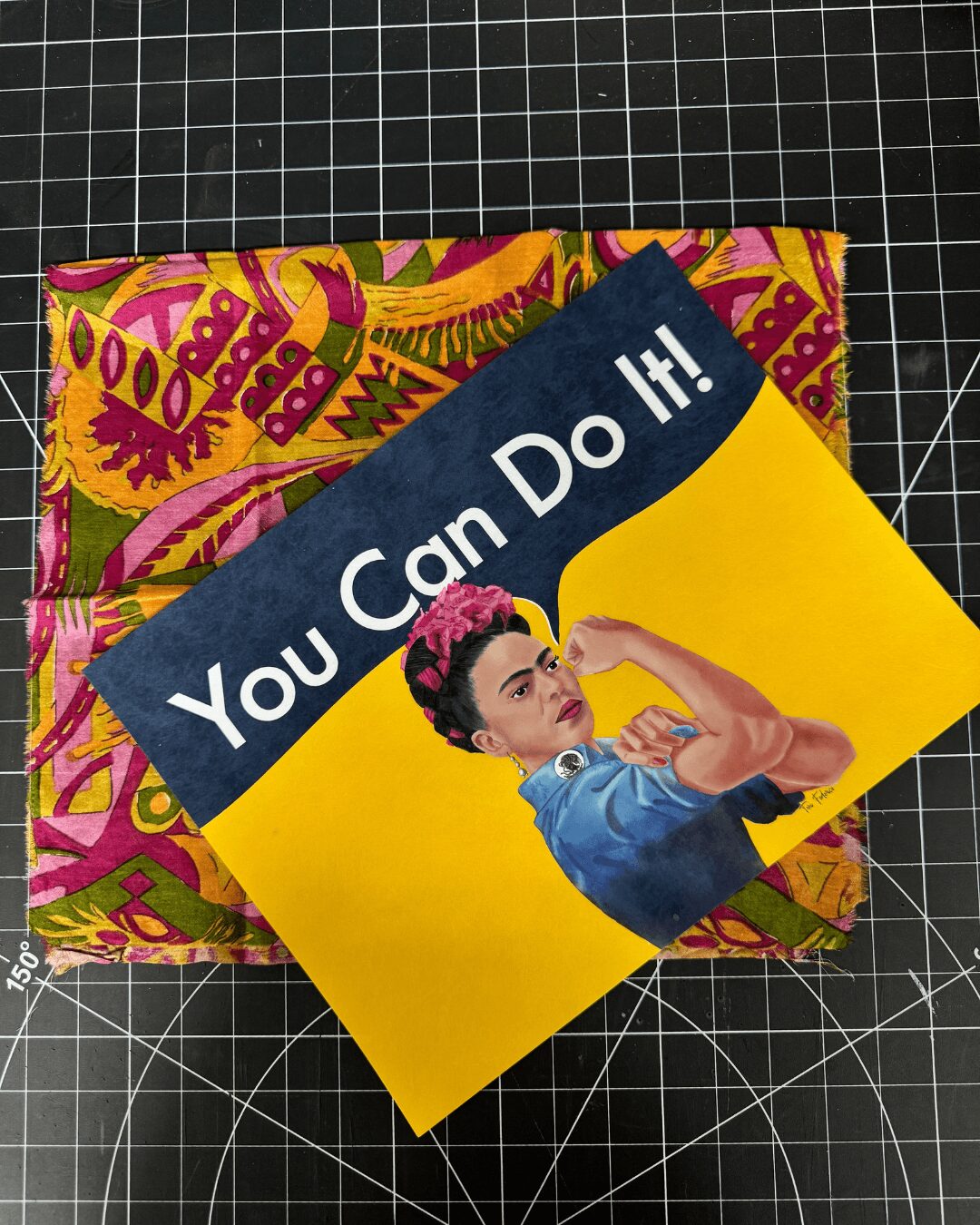 A poster with Rosie the Riveter and the text 'You Can Do It!' on a yellow background, placed on a colorful fabric.