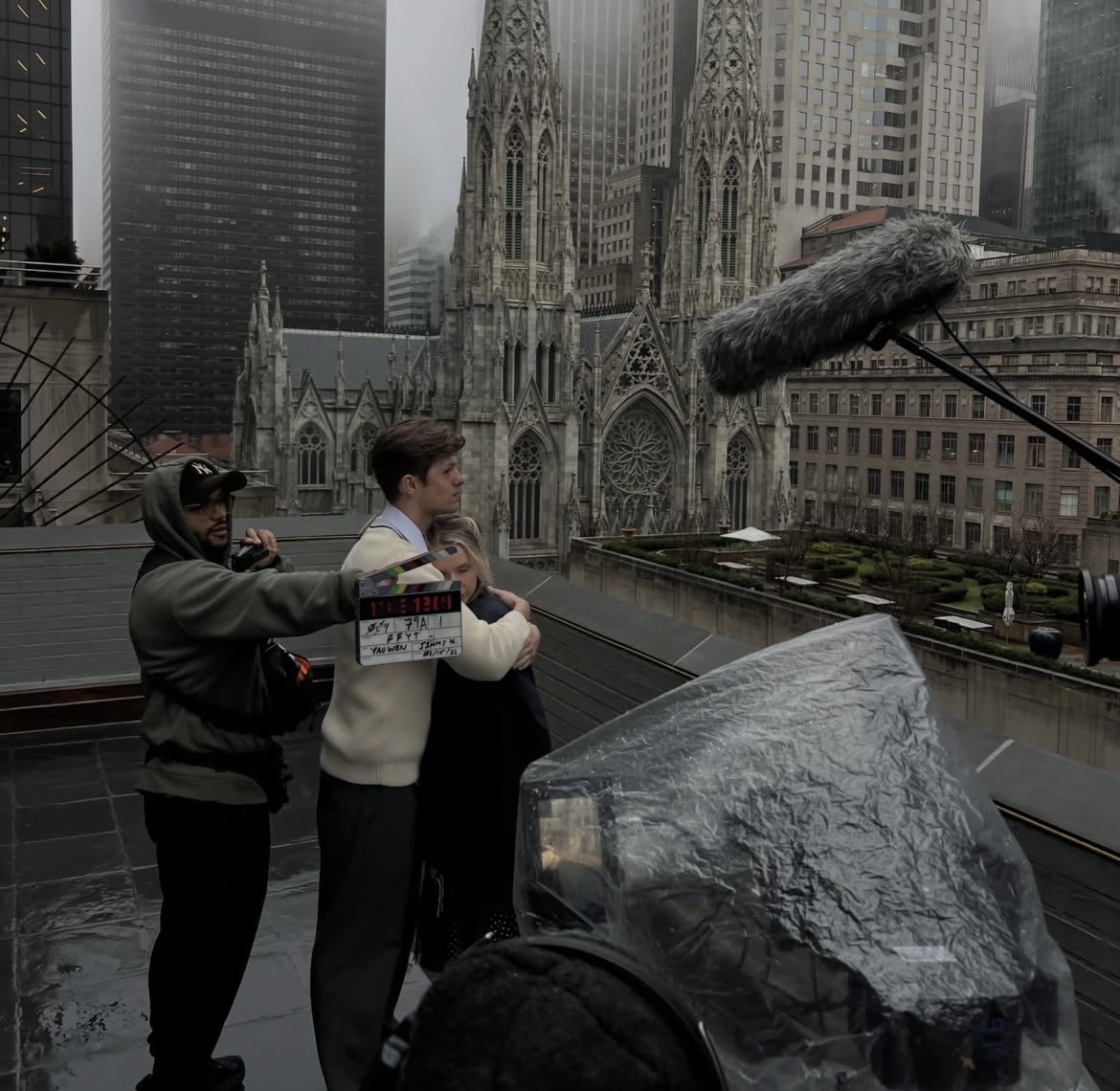 Filming crew with a microphone and camera on rooftop, cityscape with cathedral and skyscrapers in background.