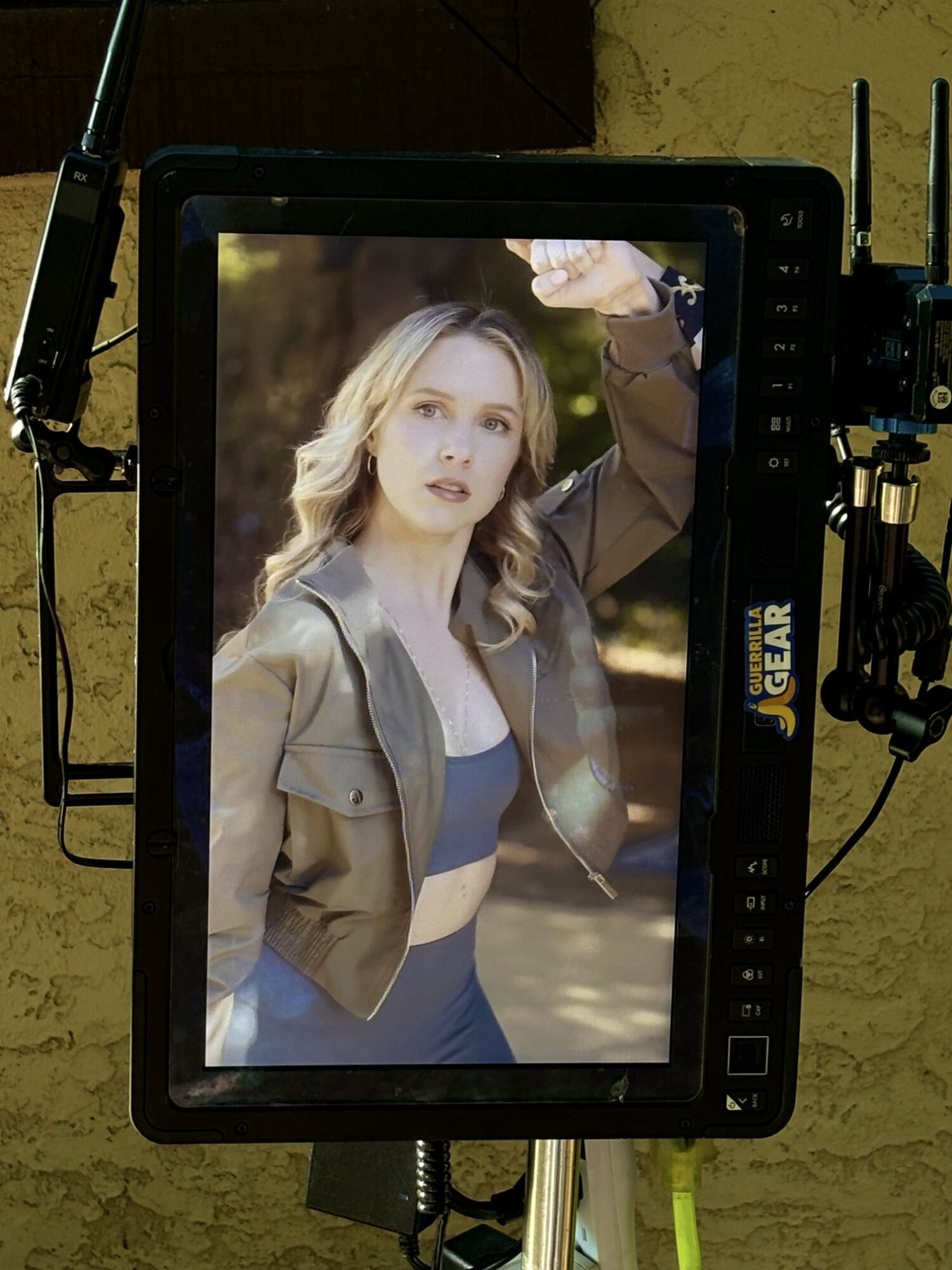 A woman with blonde hair wearing a beige jacket and blue top, standing outdoors with trees in the background.