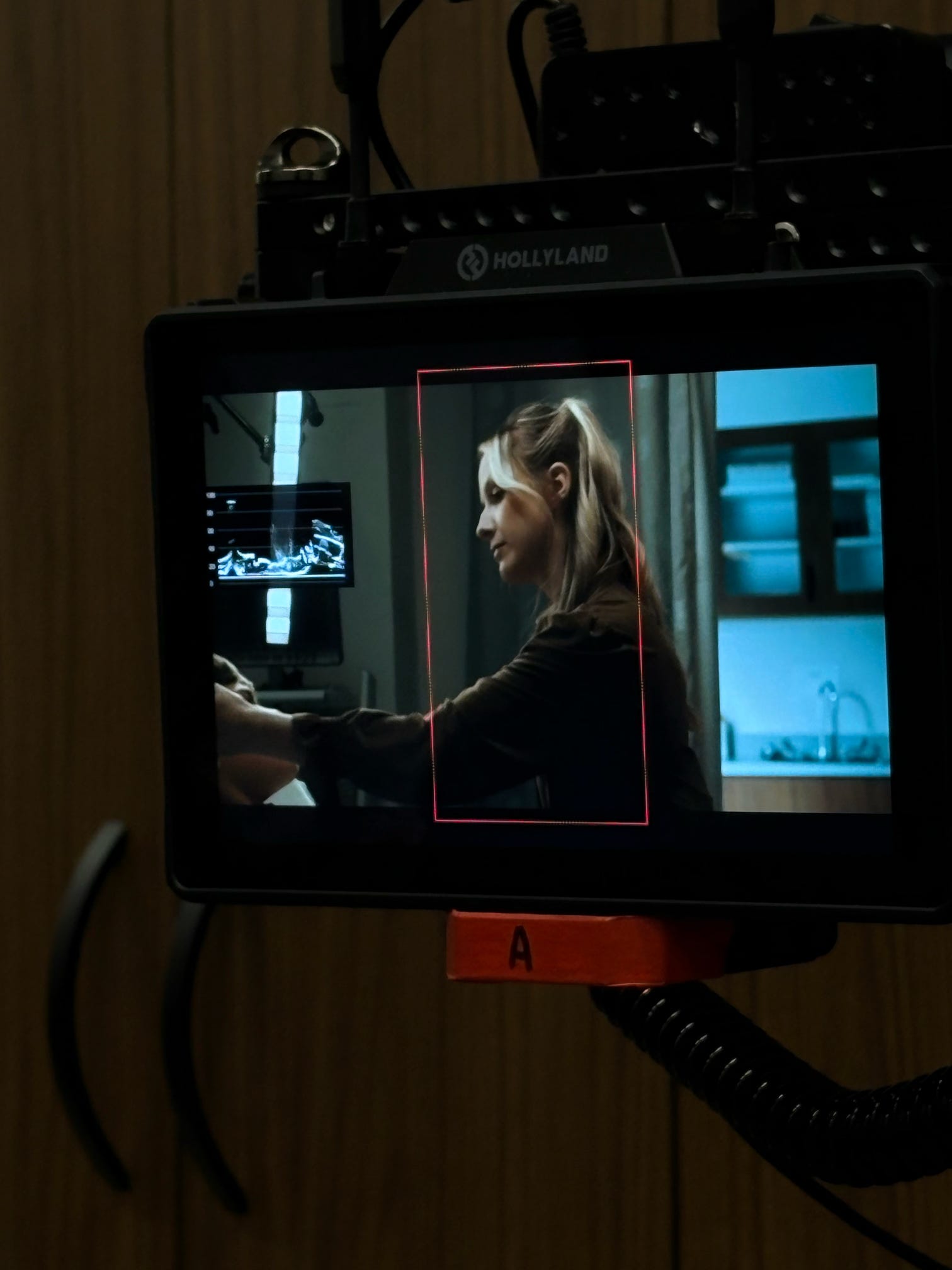 Woman with long hair in profile, sitting in a room with monitors, holding a device, illuminated by blue and white light.