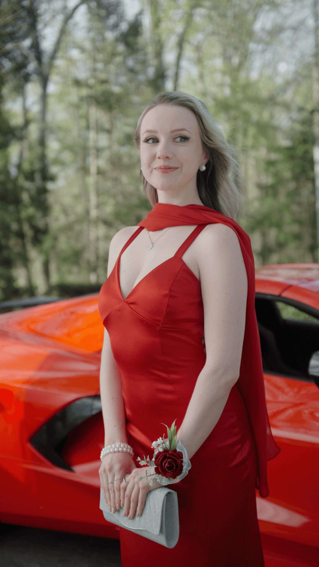Young woman in red dress holding a clutch and corsage, standing outdoors near a red sports car, with trees in background.