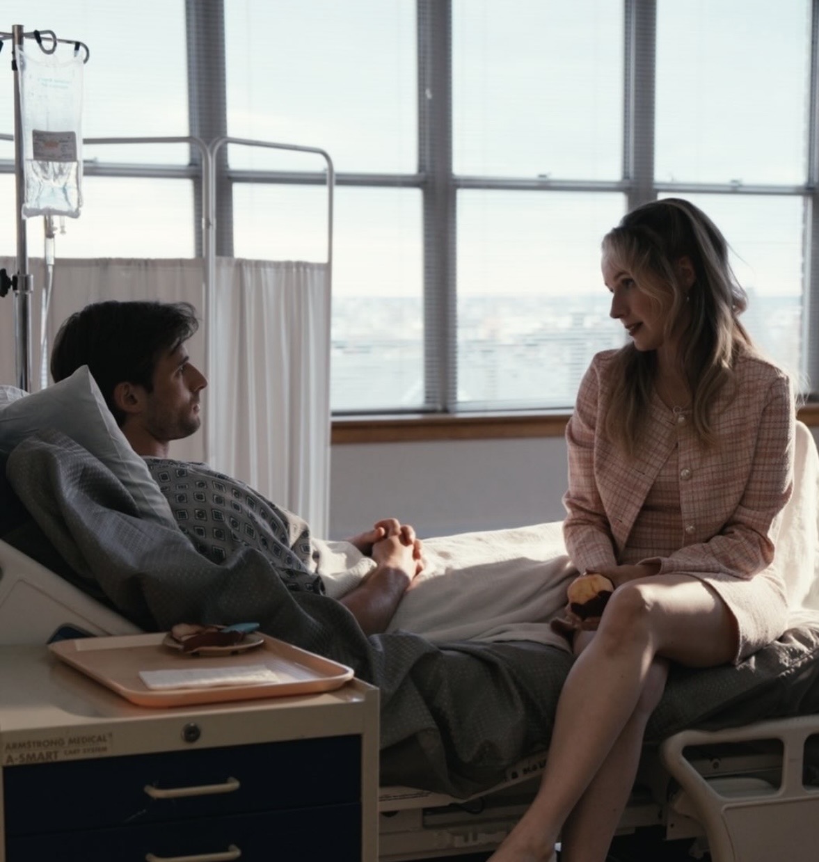 A woman and a man are talking in a hospital room with large windows, sunlight, and medical equipment nearby.